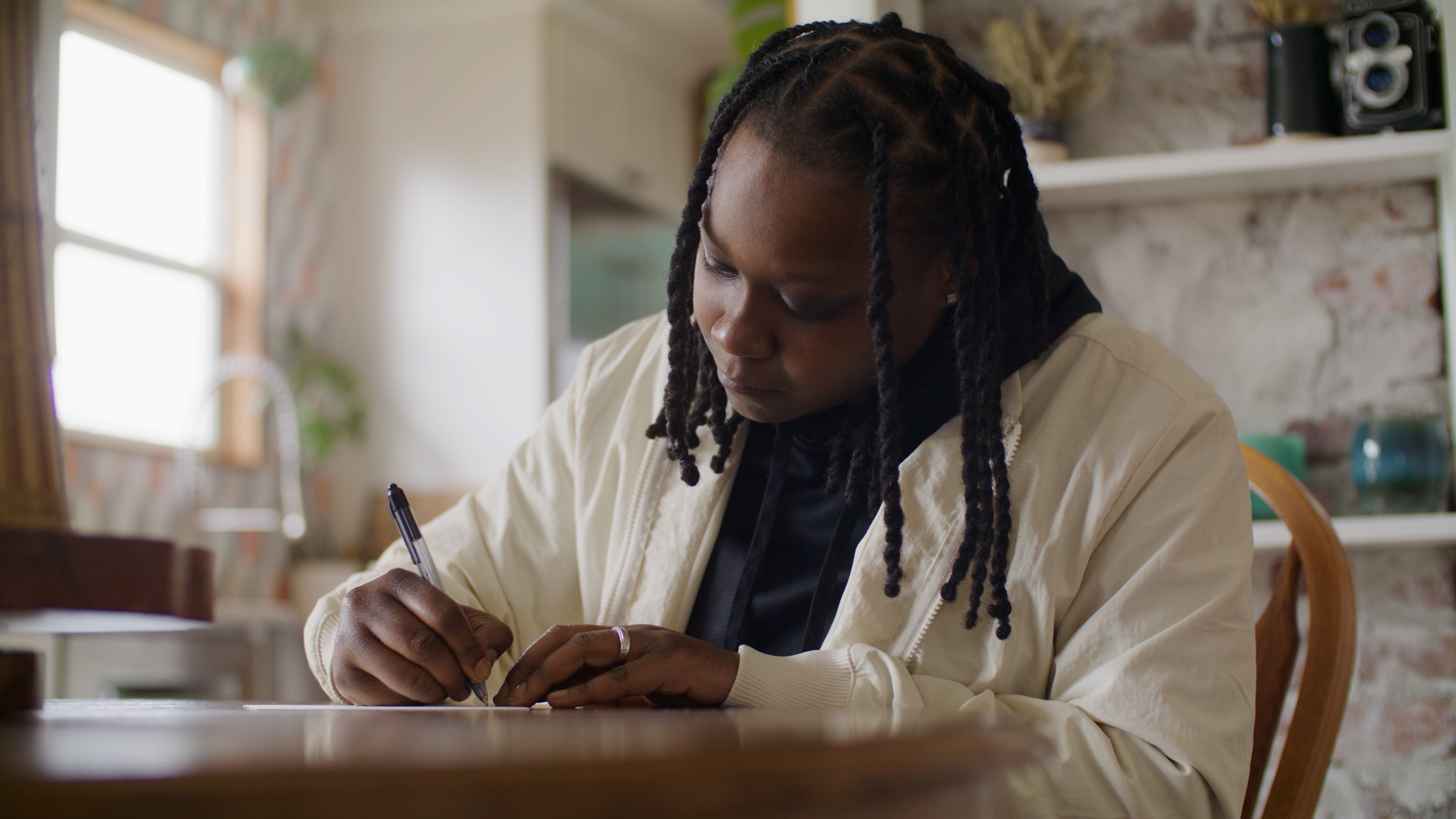 Talia writes a postcard to her younger self at a sunlit table