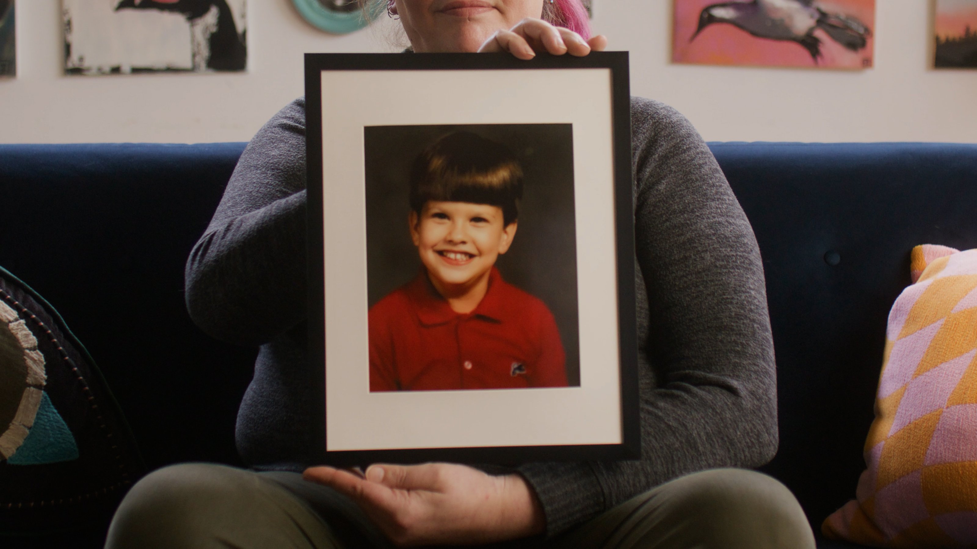 Carrie holds a framed childhood photo of herself in a room filled with colorful art