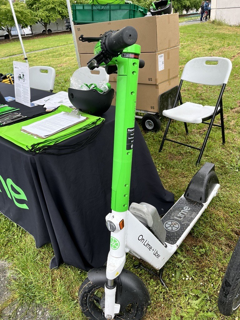 A green Lime scooter parked beside a PRR-staffed outreach table stocked with helmets and Lime tote bags at an outdoor community event