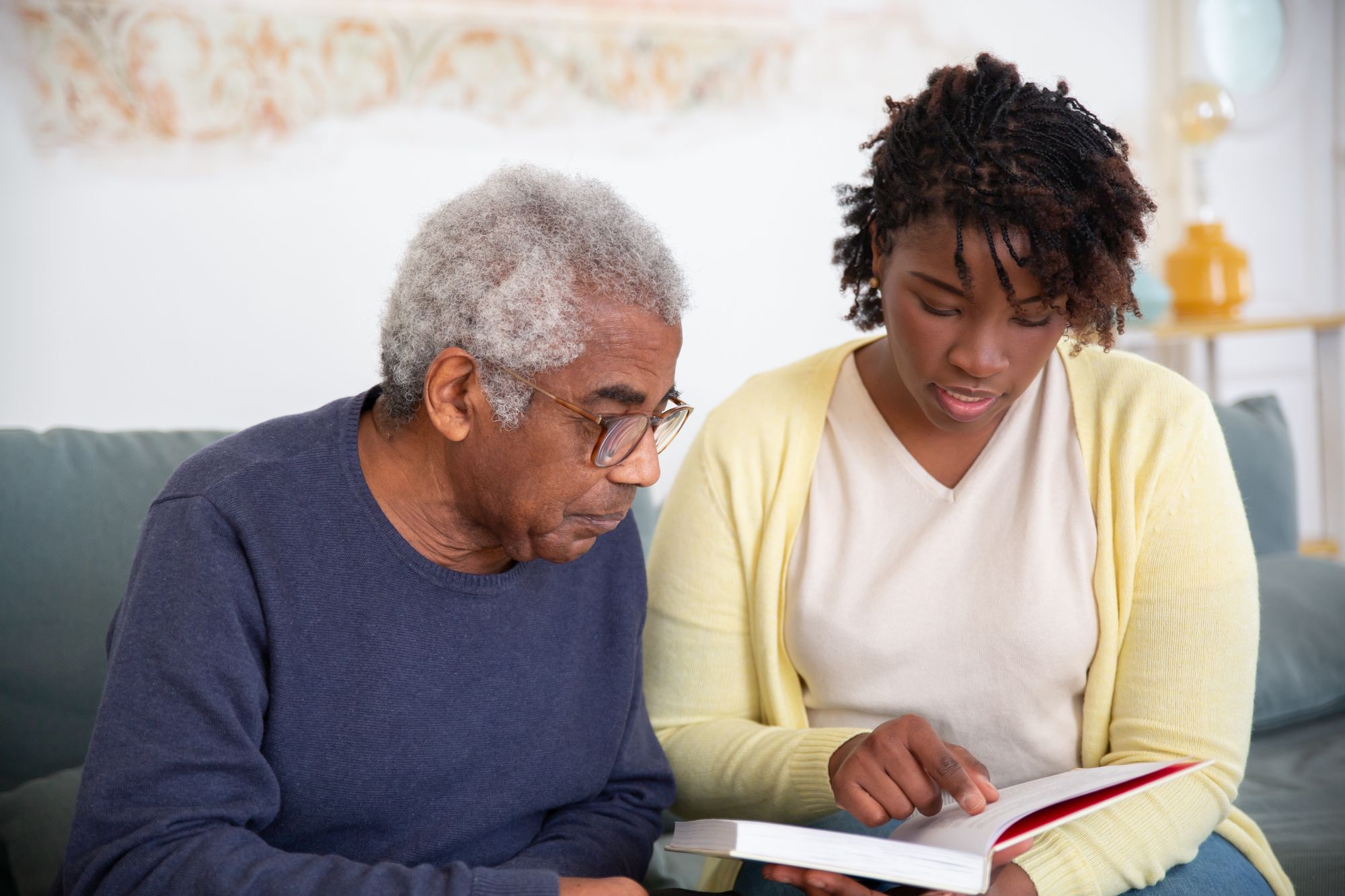 A home care worker in a yellow cardigan reads alongside an older man on the sofa in his living room