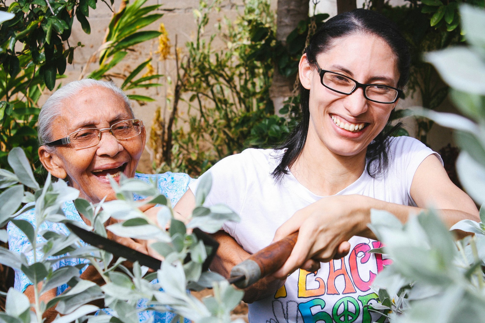 An older woman and a younger woman in t-shirts laugh together while tending plants in a garden