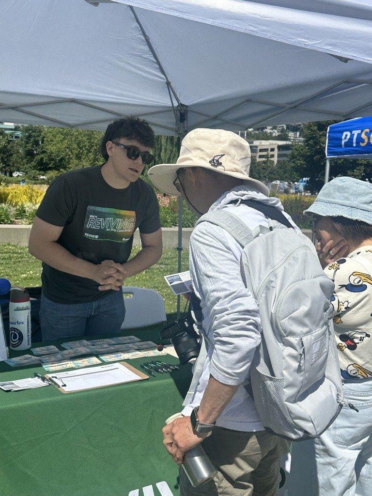 A PRR team member in a Revive I-5 shirt talks with two visitors at an outdoor project information table