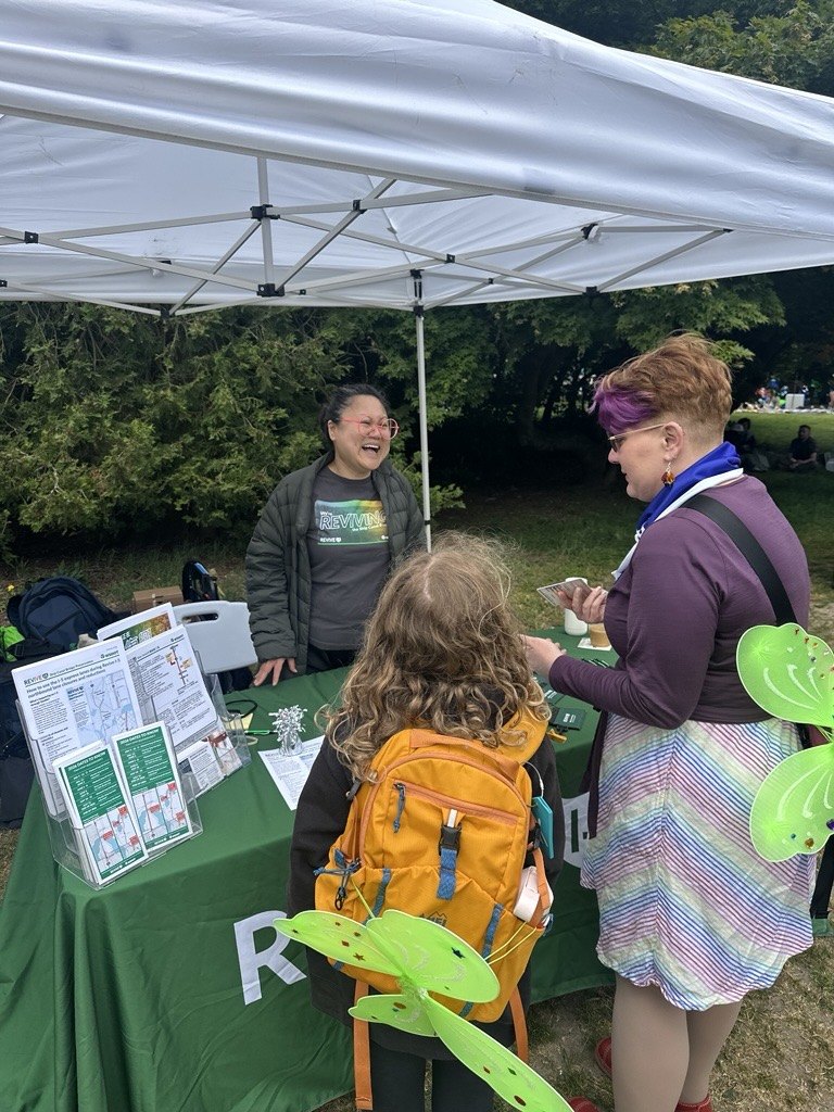 A Revive I-5 staff member hands project materials to a child and an adult at a community event table