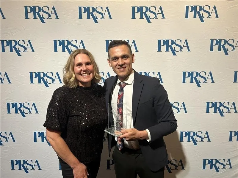 Two PRR team members holding the PRSA Silver Anvil Award of Excellence in front of a PRSA step and repeat banner.