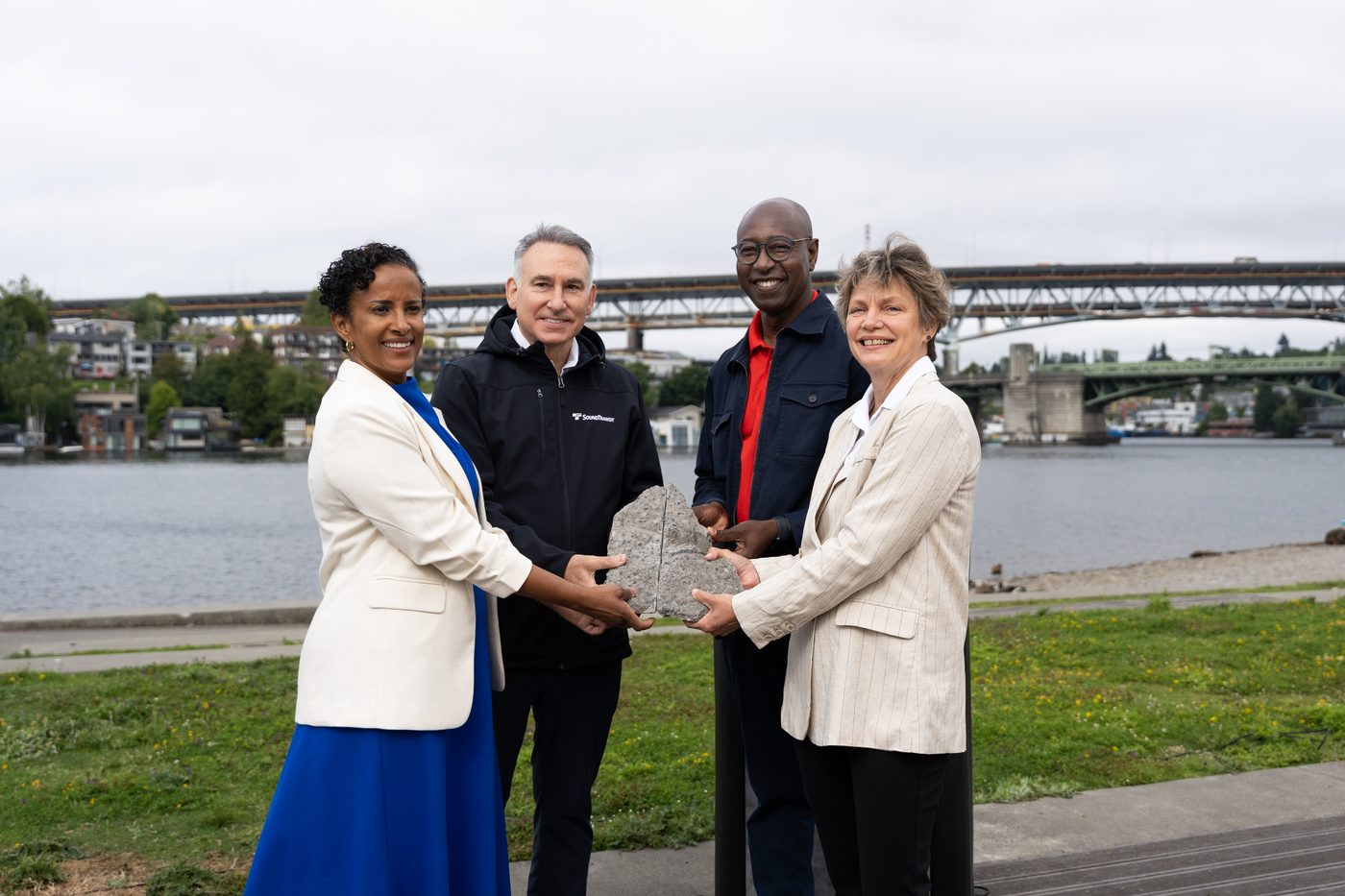 Four project partners hold a section of concrete removed from the I-5 Ship Canal Bridge, with the bridge behind them.