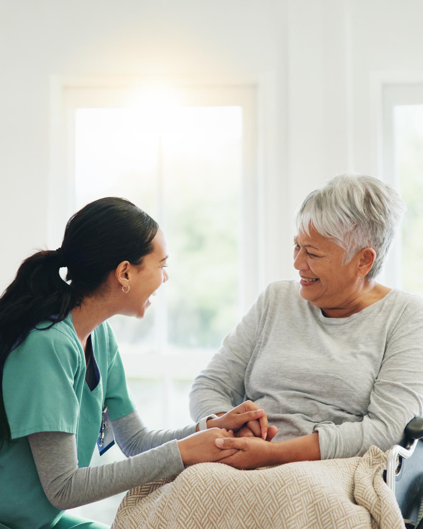 A nurse in scrubs crouching beside an older patient in a wheelchair, holding her hand as they talk.
