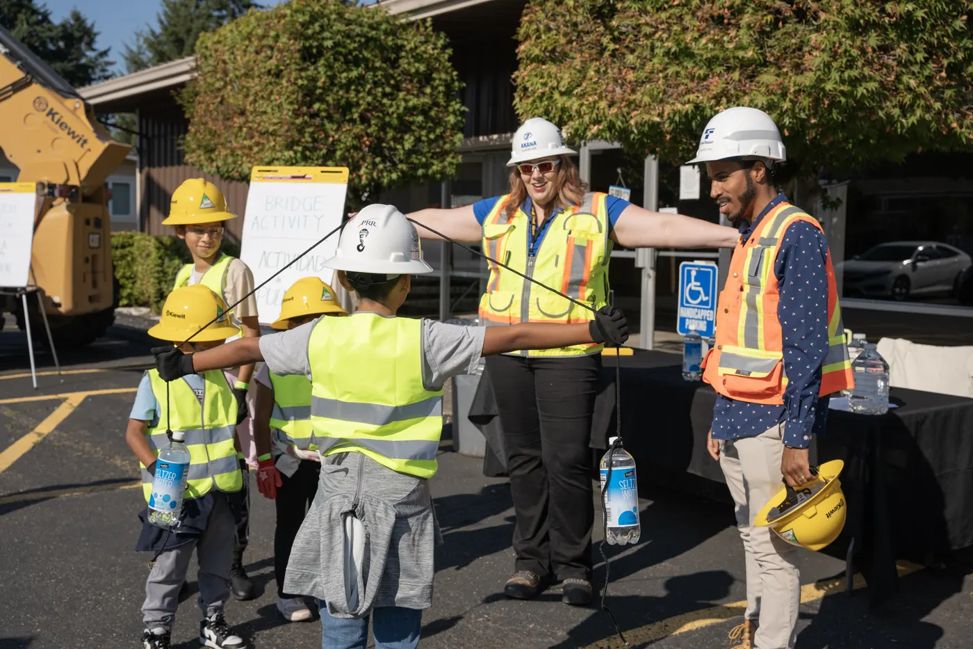A child wearing a PRR hard hat stretches out both arms holding weighted bottles during a hands-on bridge engineering activity, as two project staff look on.