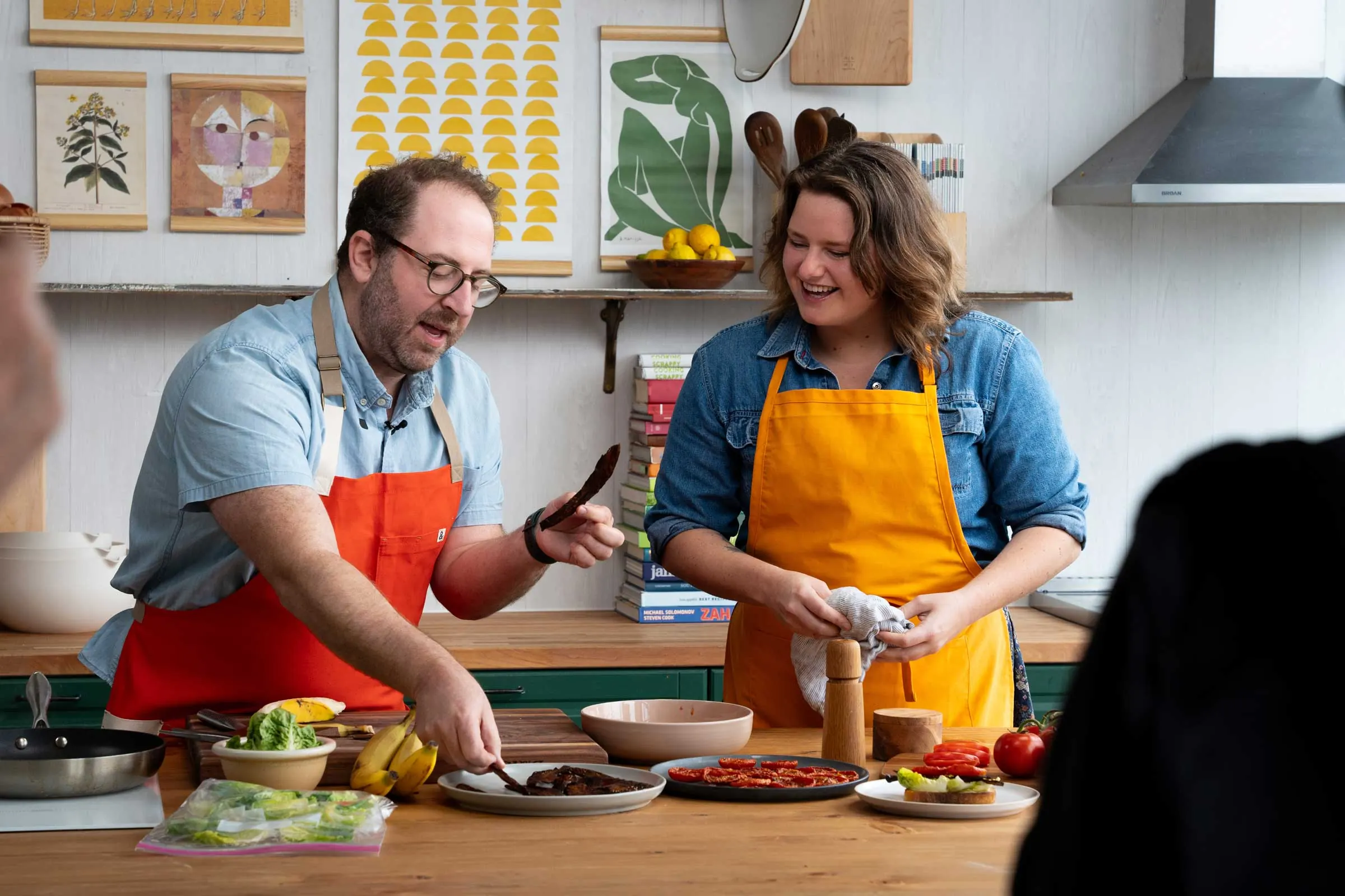 A chef in a red apron holds up a strip of roasted vegetable for a smiling co-host at a kitchen island covered with produce, prepped ingredients and leftovers during a Use Food Well video shoot