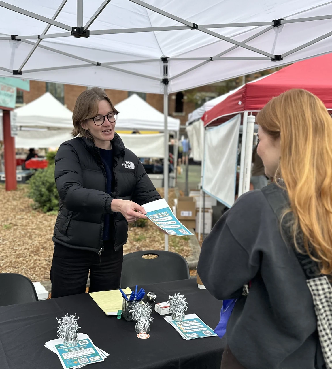 A PRR outreach staff member hands a transportation project flyer to a resident at an information table at a Portland farmers market.