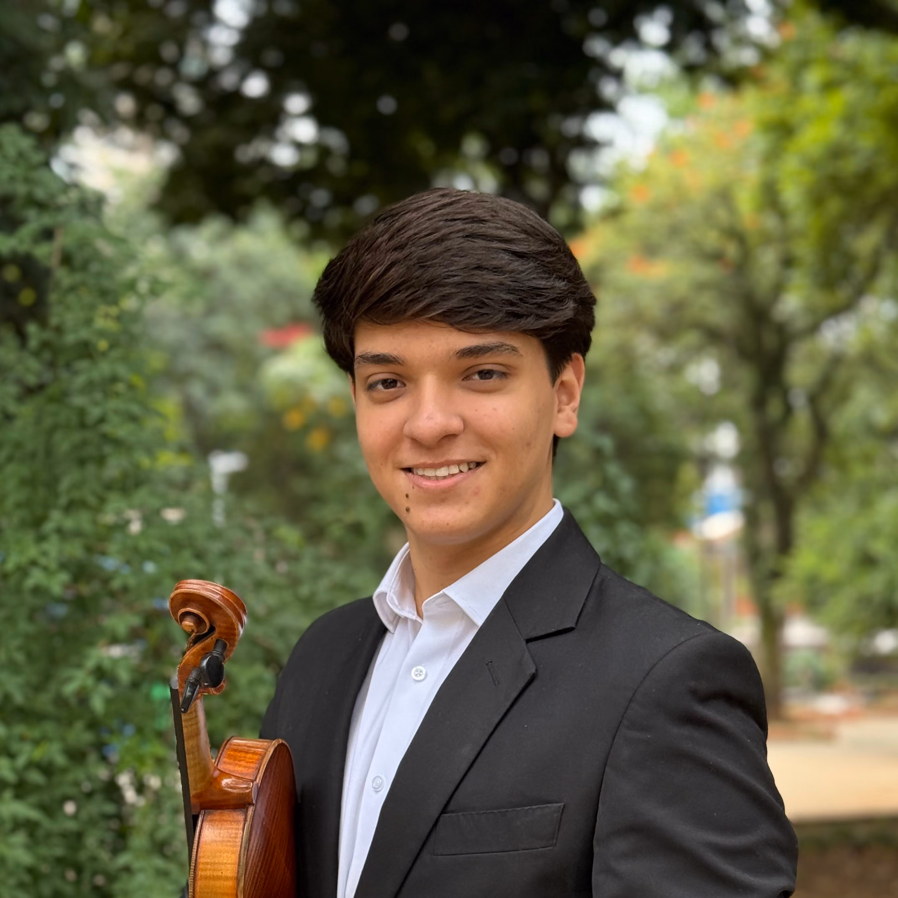 Jovem sorridente de terno preto segurando um violino em parque arborizado.