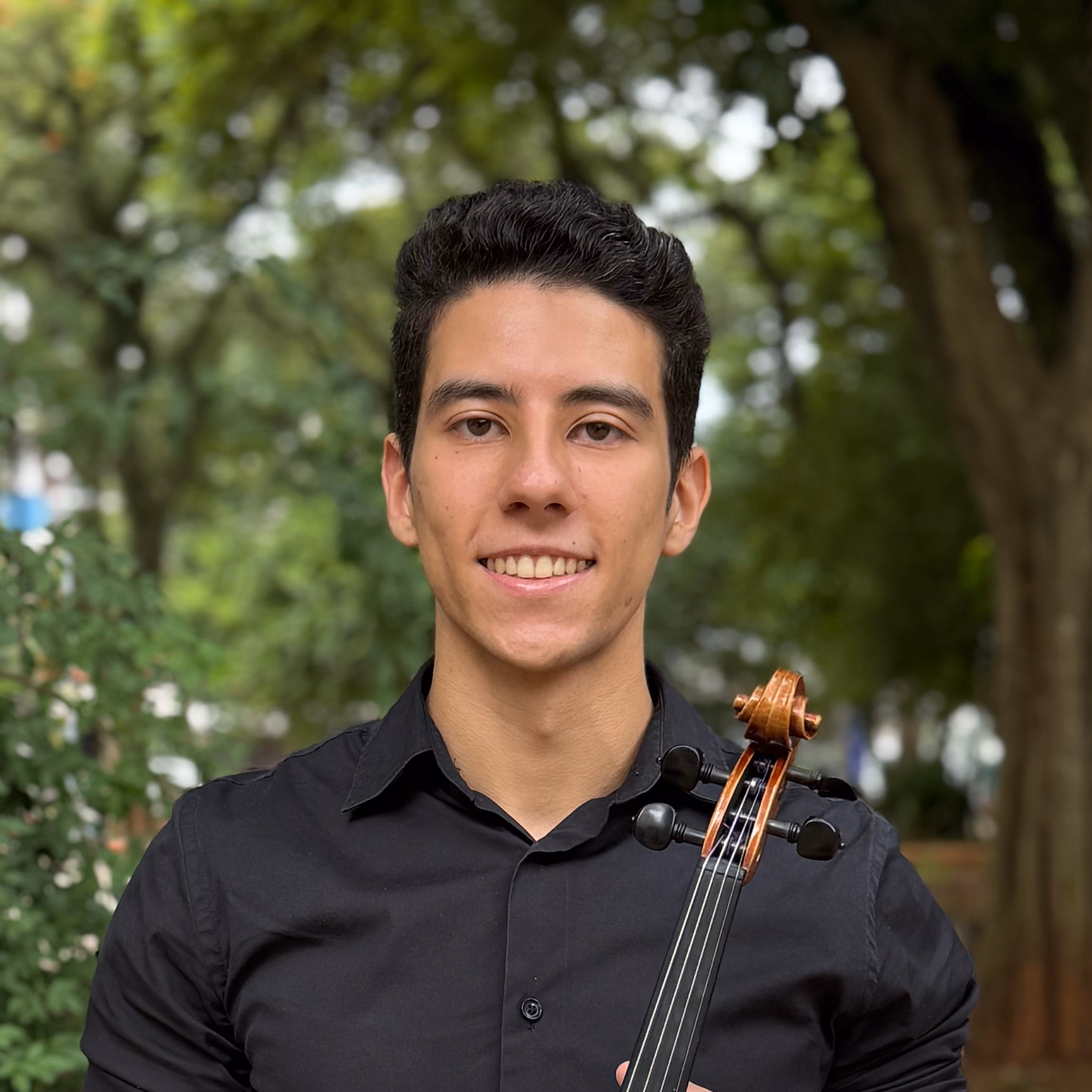 Jovem sorridente com camisa preta segurando um violino ao ar livre com árvores ao fundo.