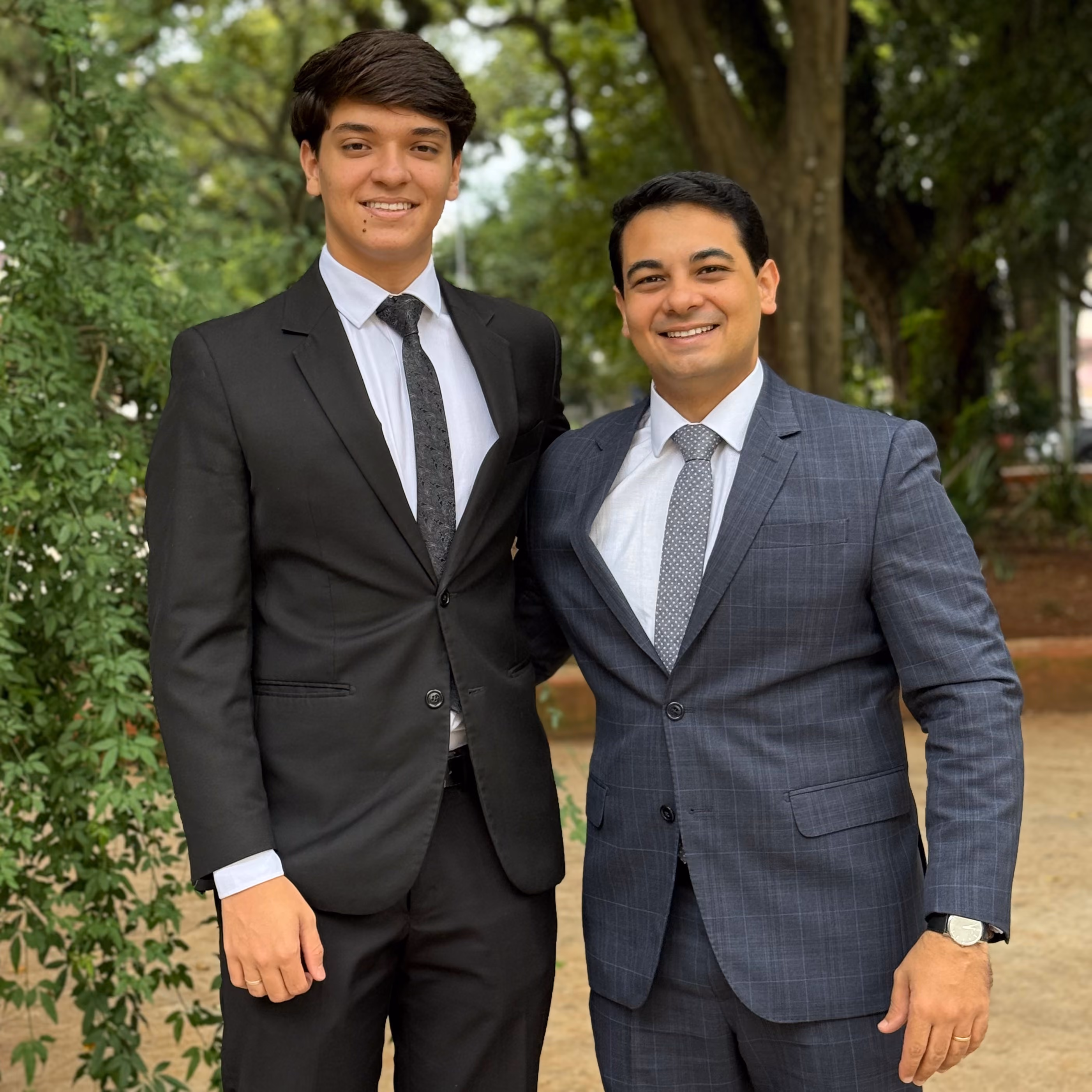 Dois homens sorrindo usando ternos formais em uma área externa com árvores ao fundo.