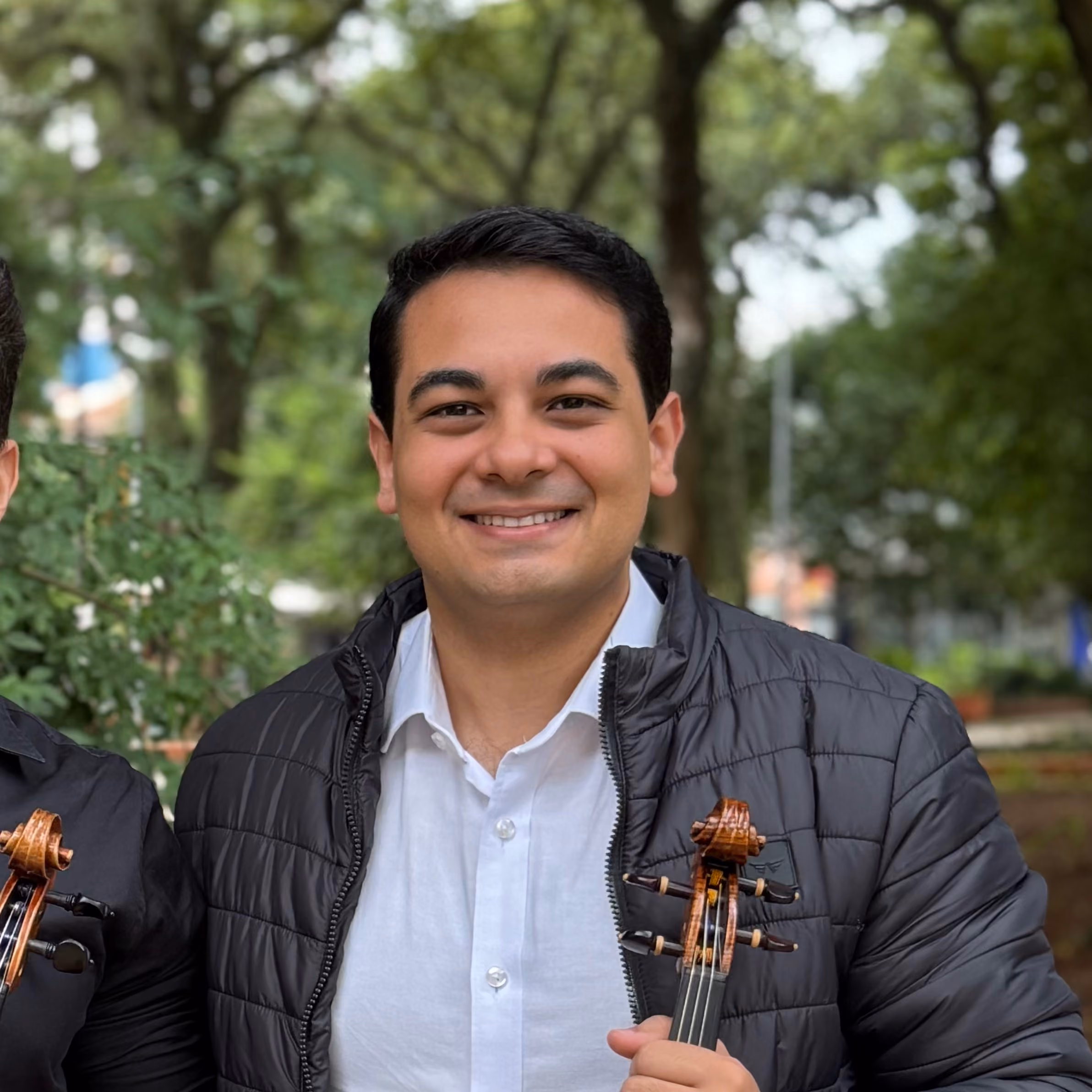 Homem sorridente segurando um violino, vestindo camisa branca e jaqueta preta ao ar livre.
