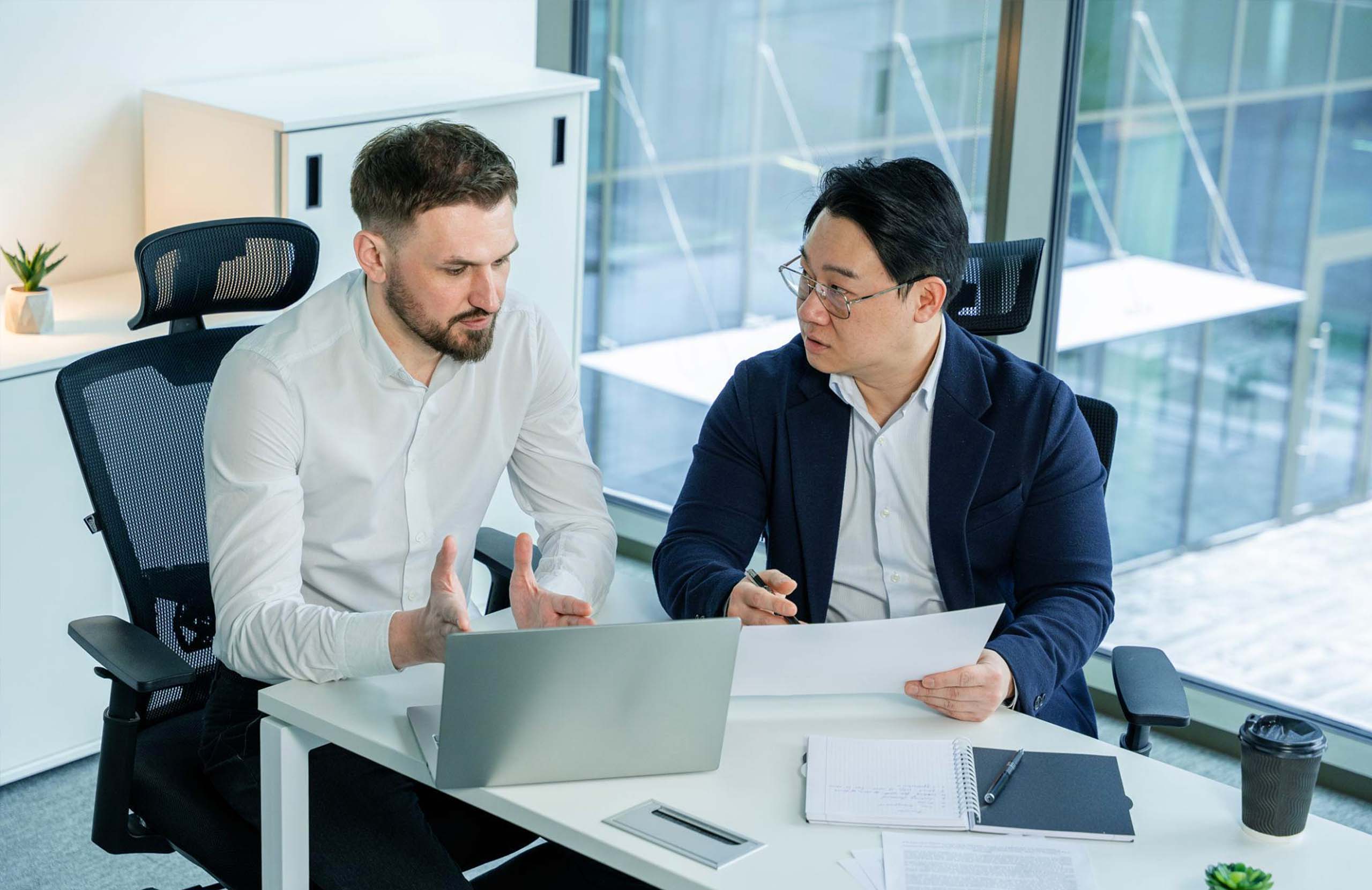 Two professionals discussing documents and reviewing information on a laptop during a meeting in a modern office.
