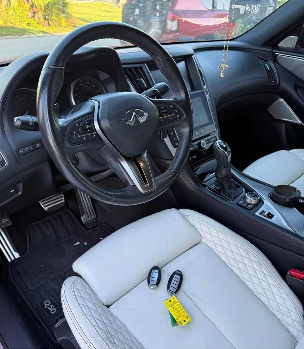 Interior of Infiniti Q50 showing steering wheel, white quilted leather seat, gear shift, and two key fobs on seat.