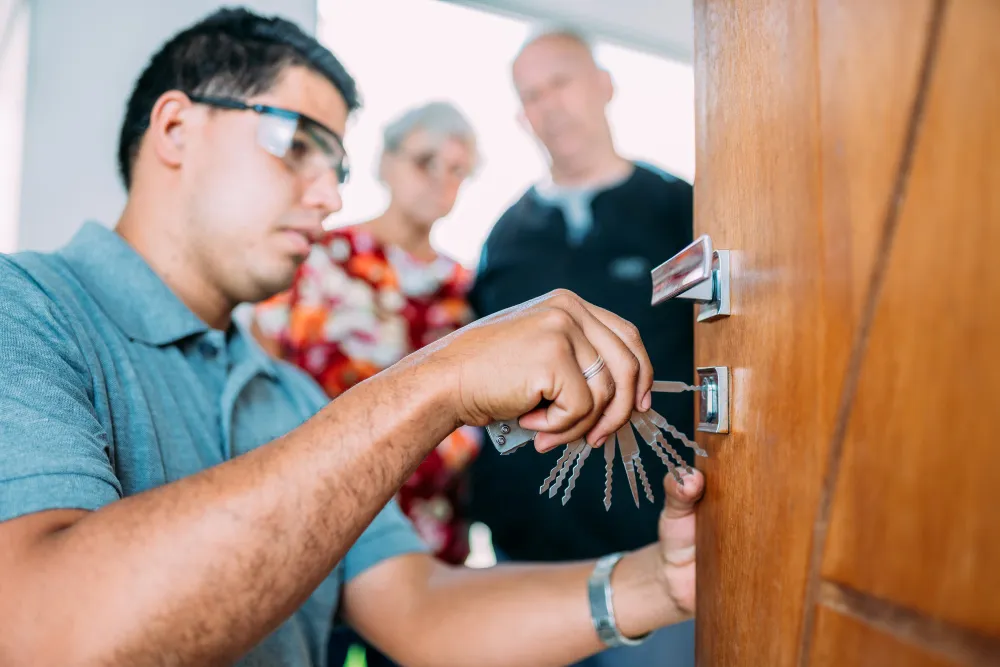Locksmith using a set of lock picks on a wooden door, with two people watching in background.
