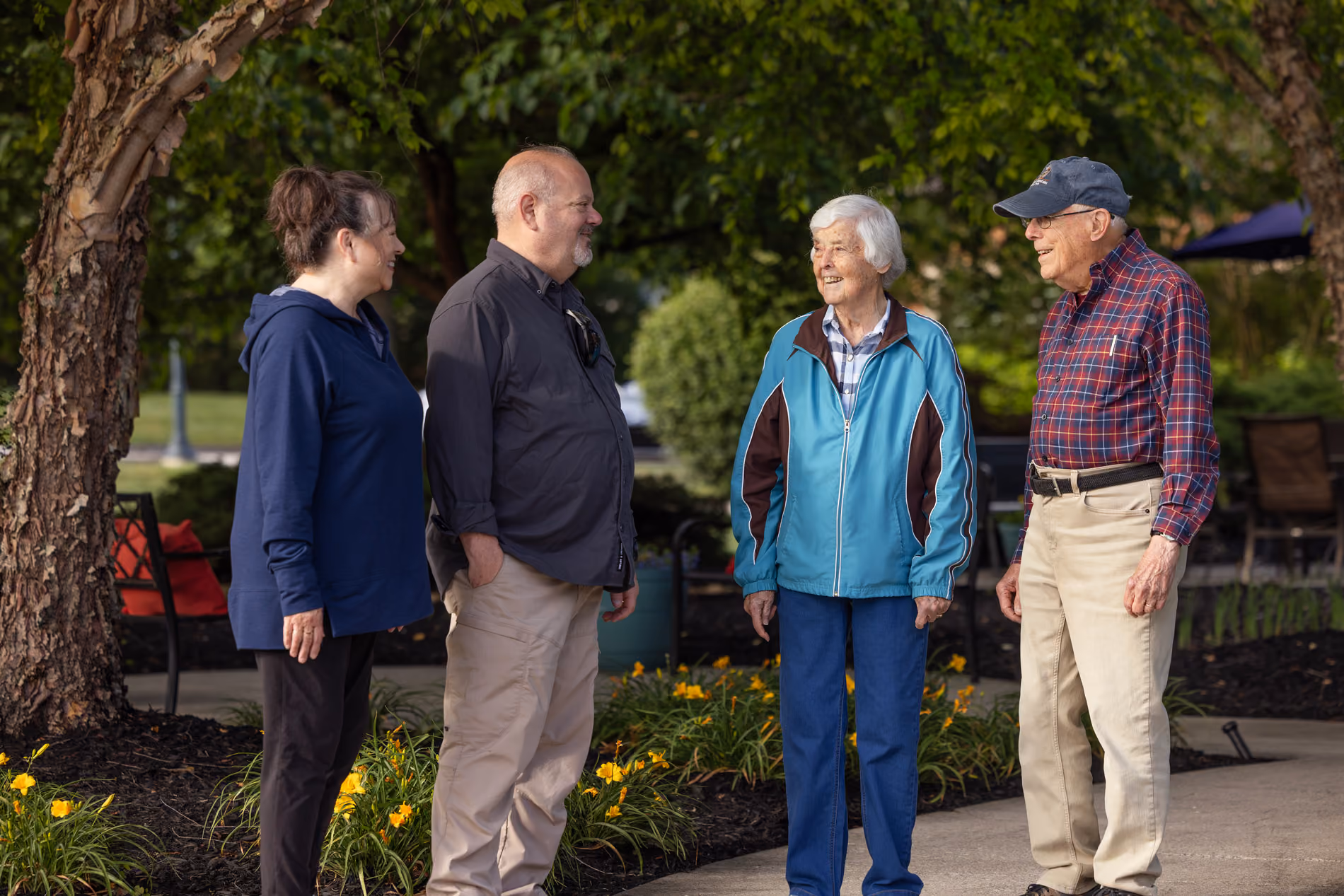 Two elderly and two middle-aged people standing outdoors in a garden, smiling and talking.