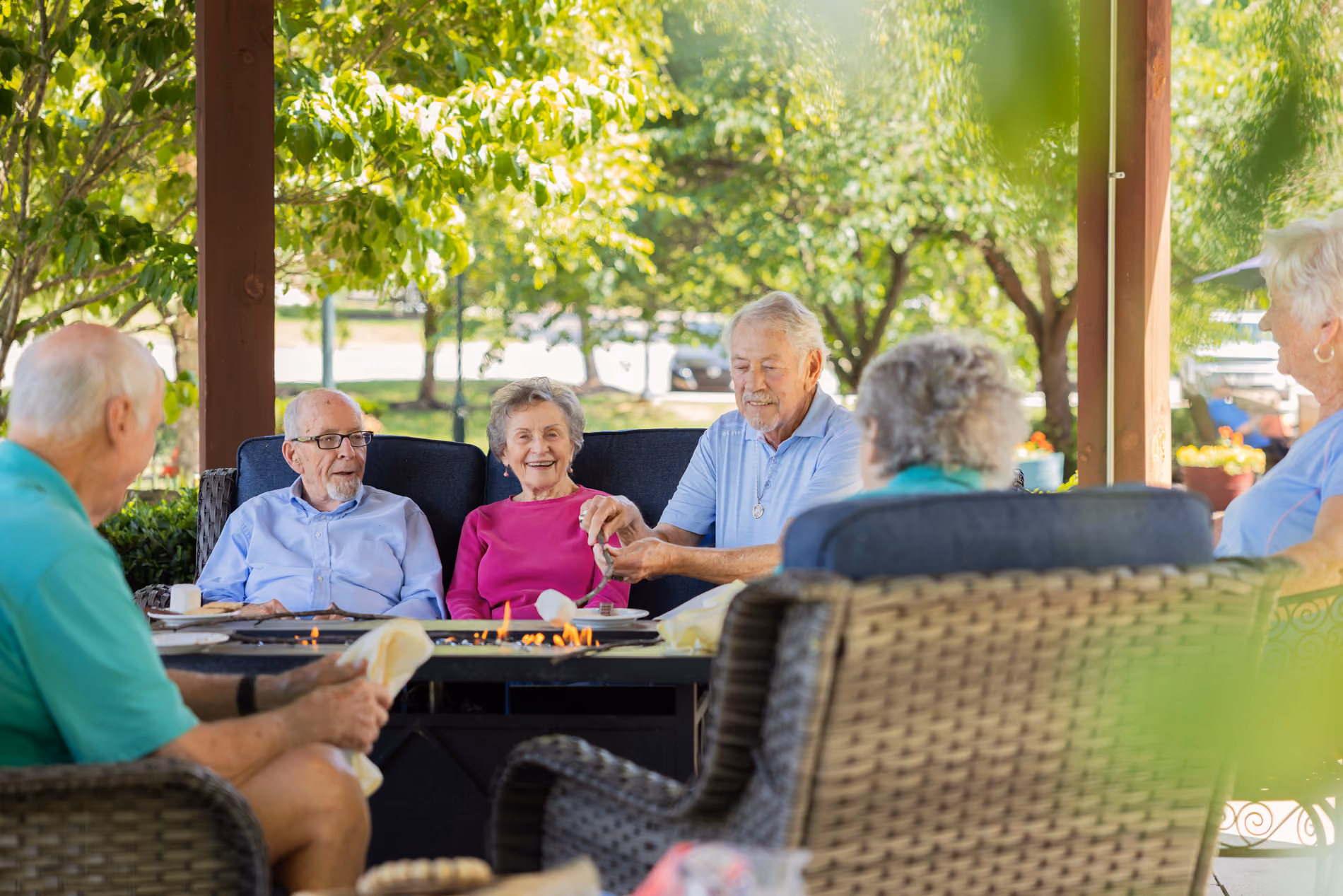 Group of elderly people sitting outdoors around a firepit, with one man roasting marshmallows.