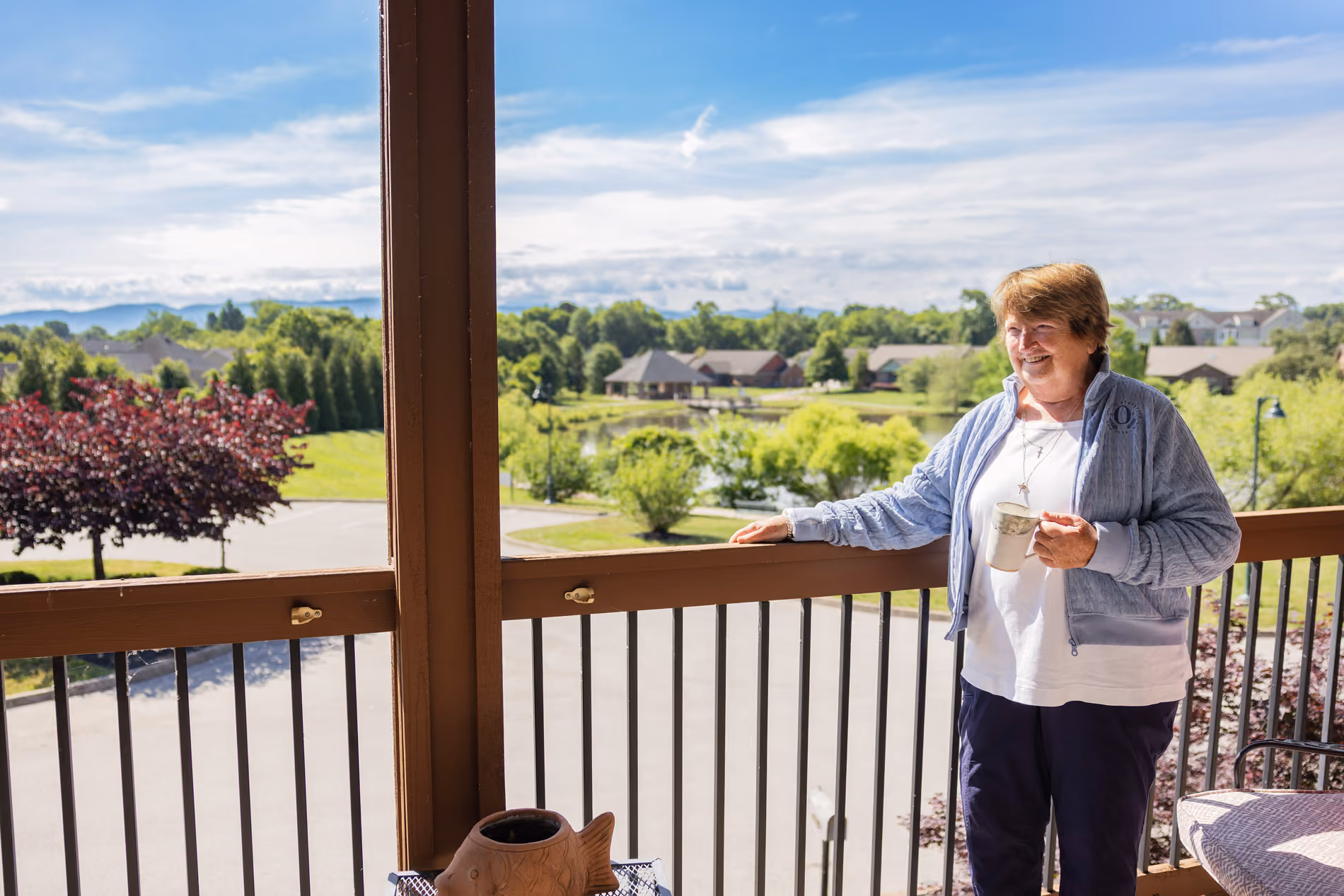 Smiling older woman holding a mug on a balcony overlooking trees, houses, and a pond on a sunny day.