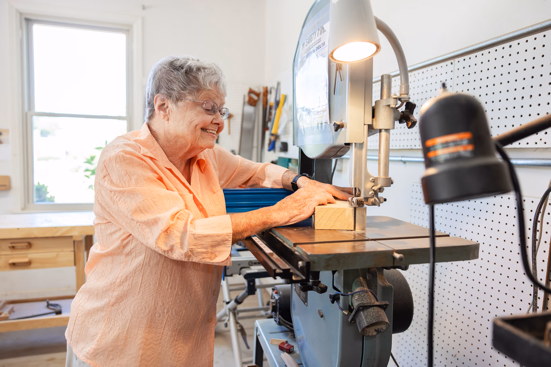 Elderly woman using a band saw to cut a piece of wood in a bright workshop.