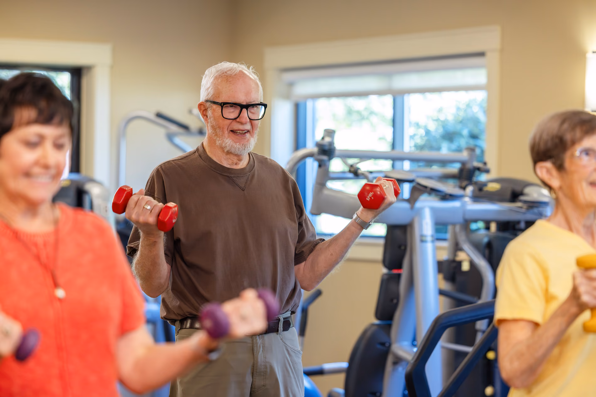 Senior man lifting red dumbbells in a gym with two women also exercising nearby.
