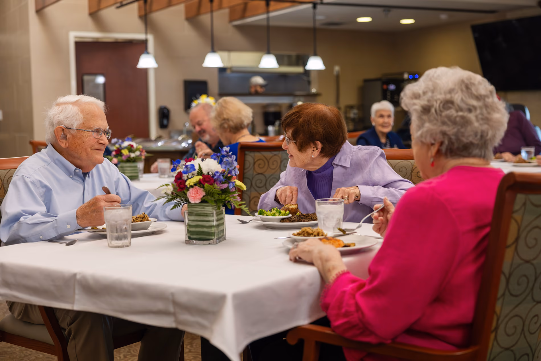 Elderly people sitting at a dining table with food and flowers, enjoying a meal and conversation.