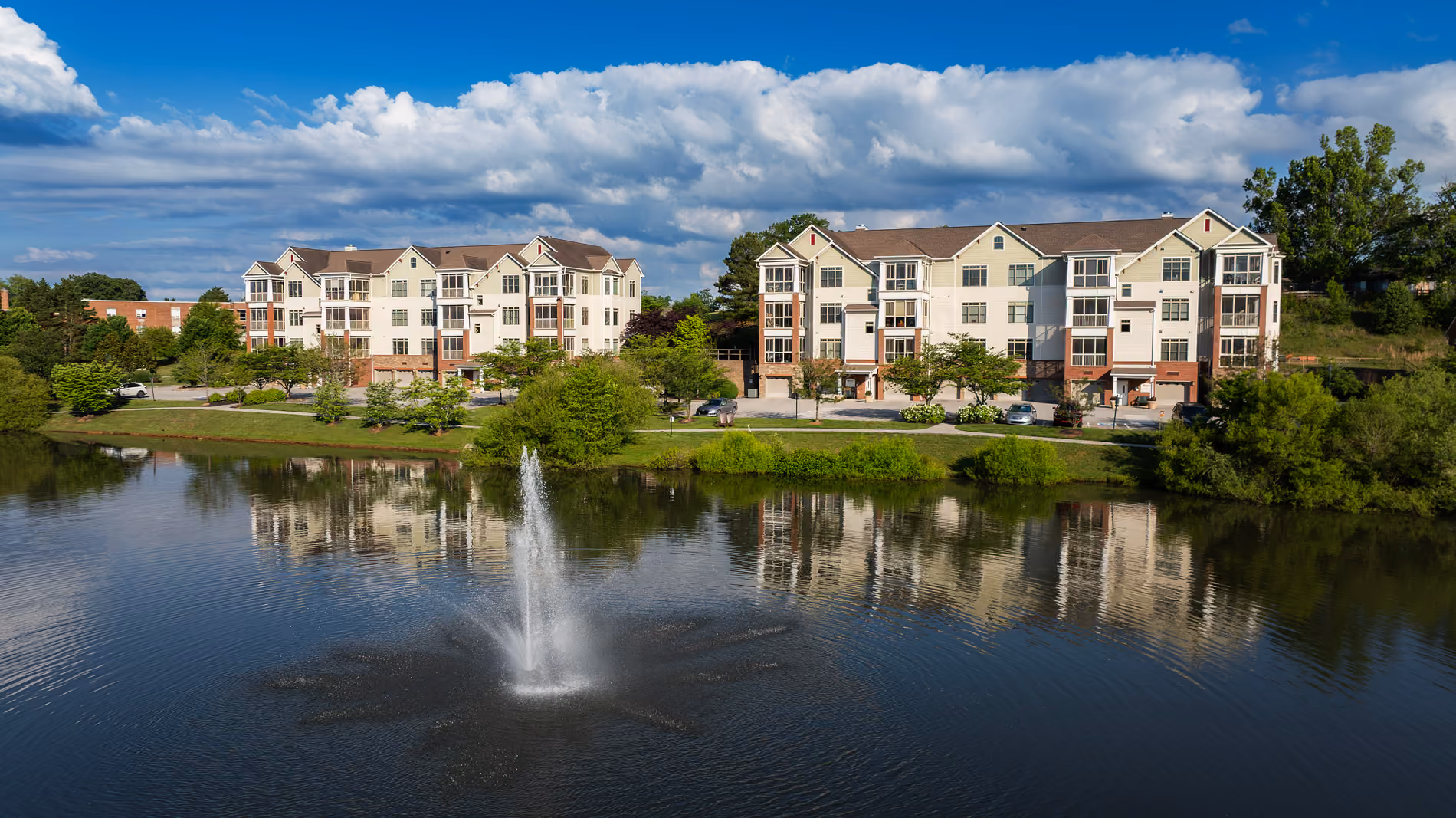 Residential buildings reflected in pond with a central water fountain under a blue sky with clouds.