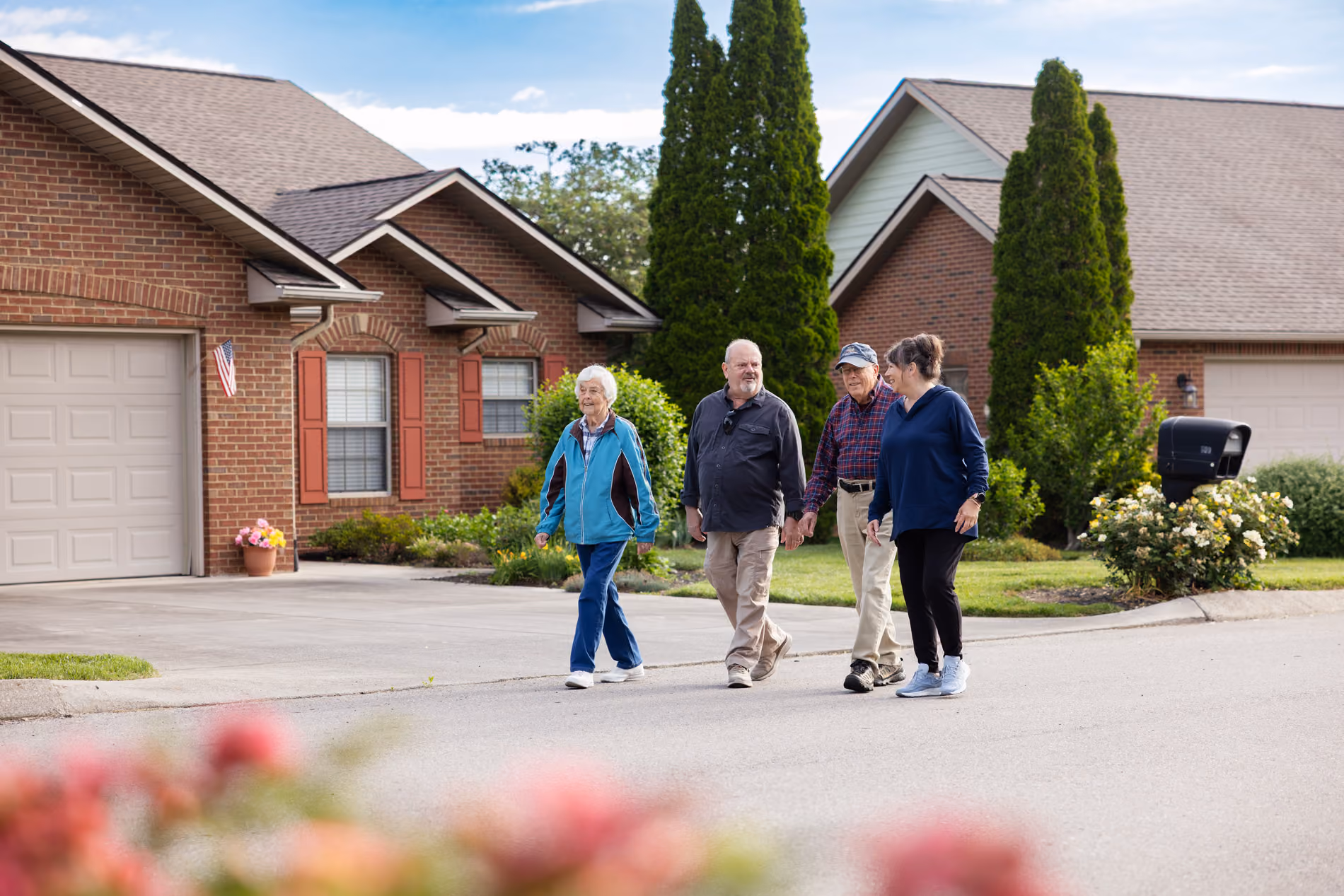 Four older adults walking and chatting on a residential street with brick houses and greenery.