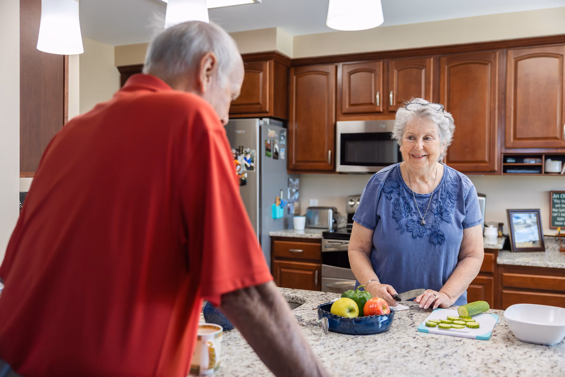 Elderly woman slicing cucumber in kitchen while smiling at elderly man in red shirt.
