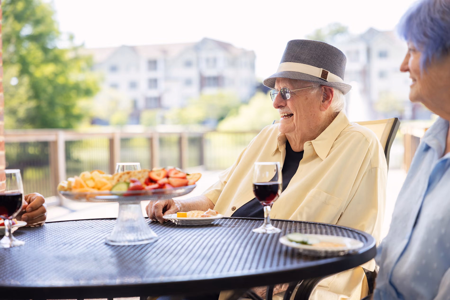 Elderly man smiling outdoors at a table with wine and a plate of snacks, talking to others.