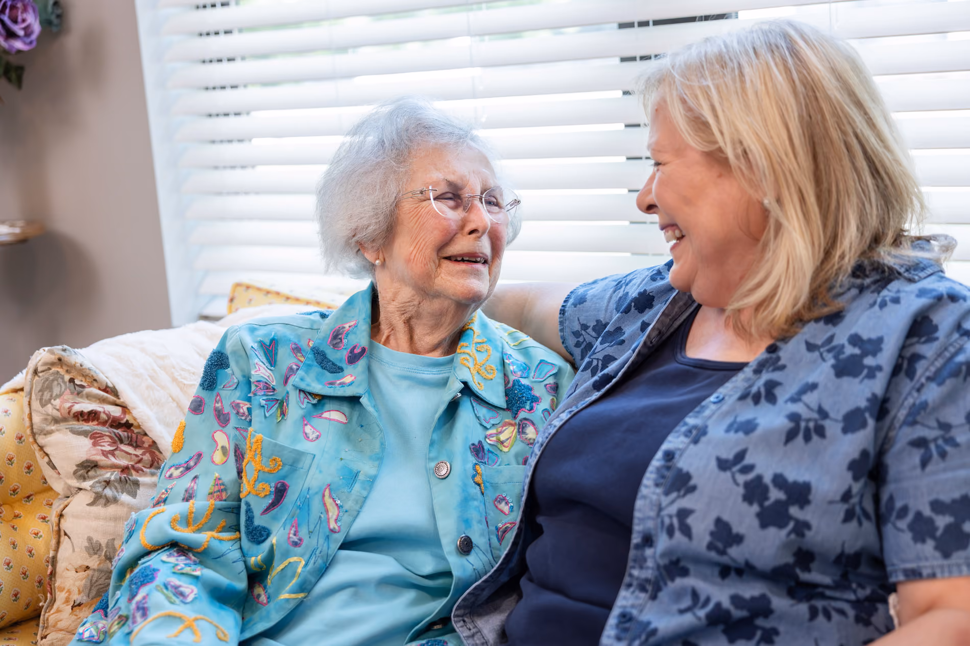 Two women sitting on a couch, smiling warmly at each other in a cozy room with white blinds.