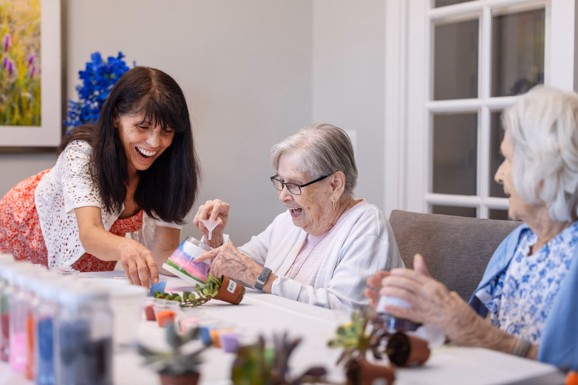 Woman helping two elderly women with an arts and crafts project at a table with plants and paints.