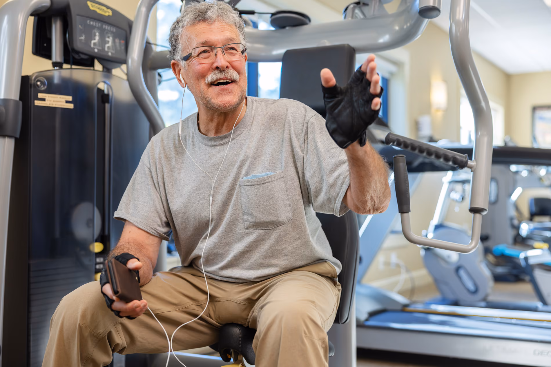 Older man wearing gloves and earbuds, sitting on gym equipment and holding a phone, smiling and gesturing.