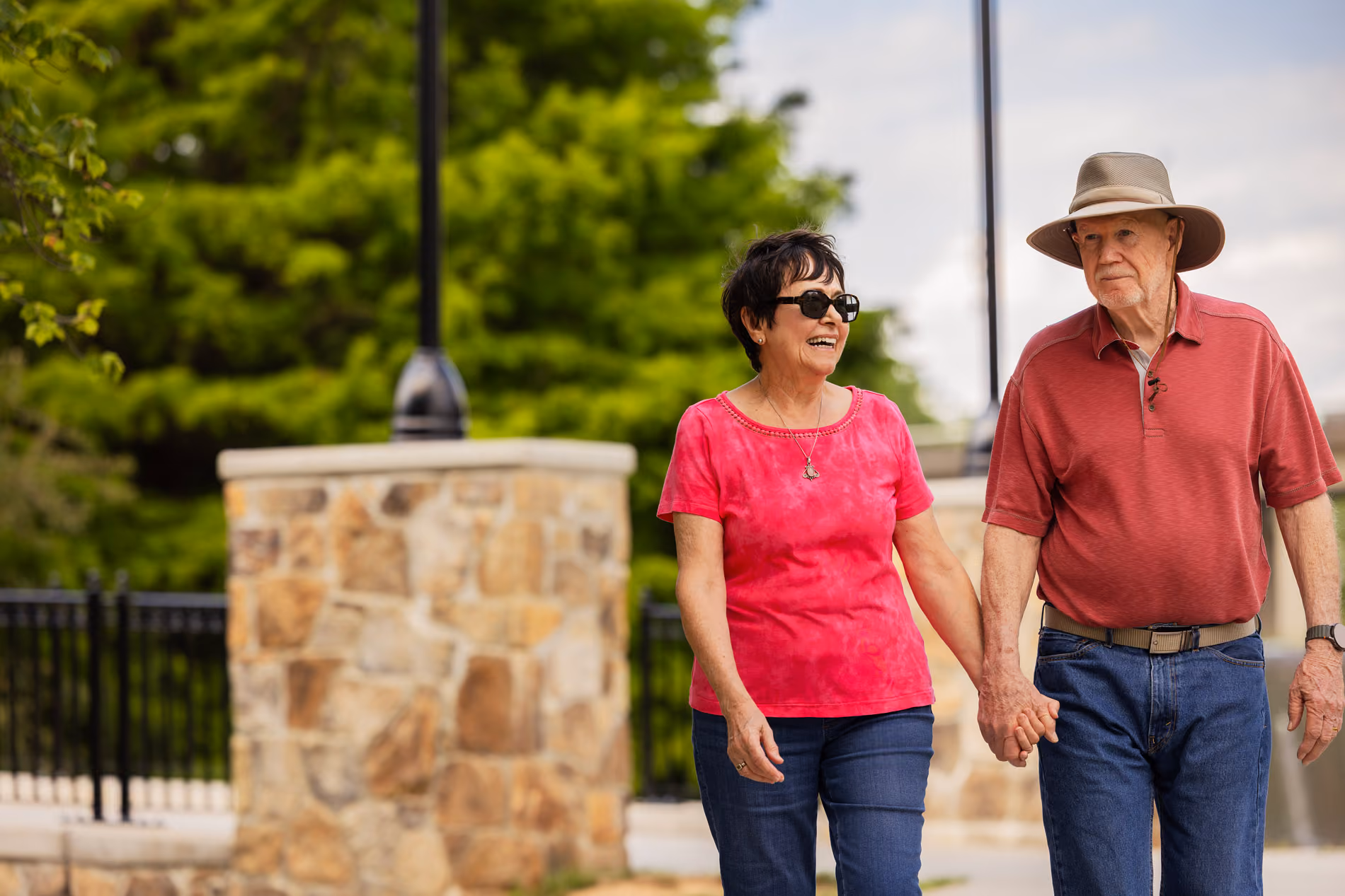 Older couple holding hands walking outdoors, woman smiling and man wearing a sun hat.
