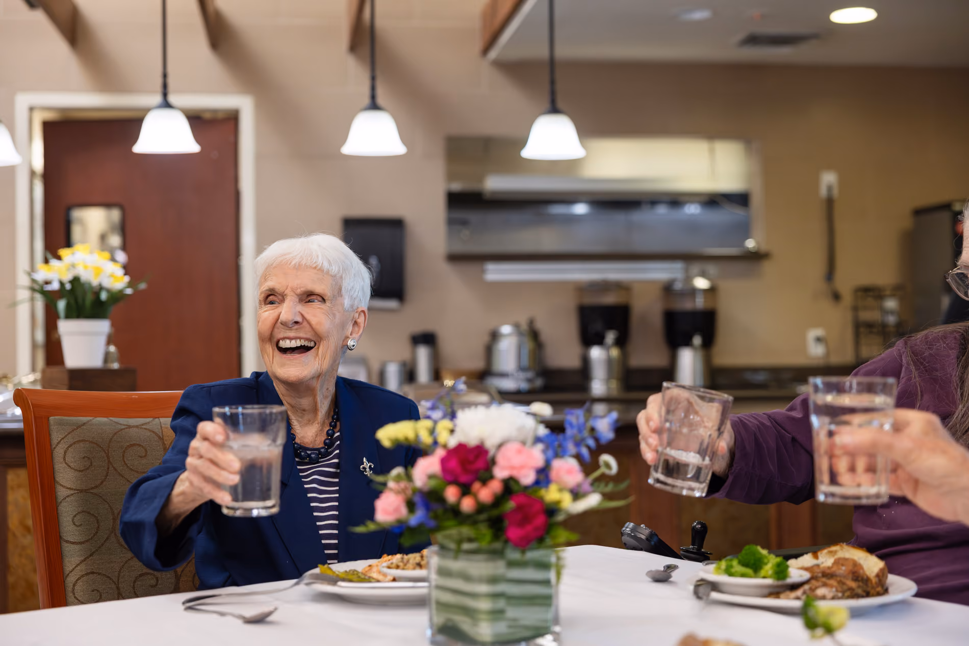 Elderly woman smiling and raising a glass at a dining table with colorful flowers in the center.