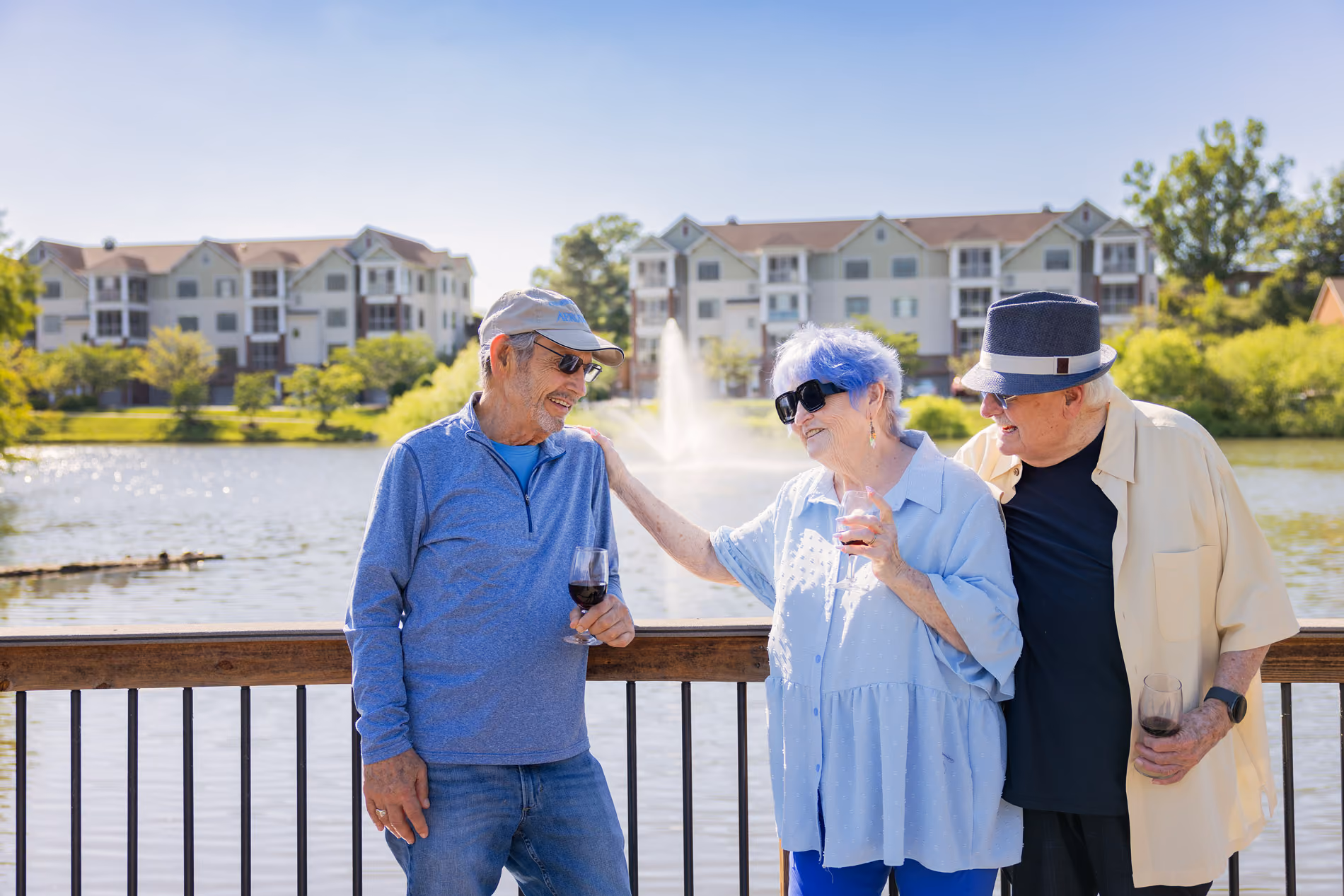 Three elderly friends with sunglasses and hats enjoying wine by a lake with apartment buildings and fountain behind.