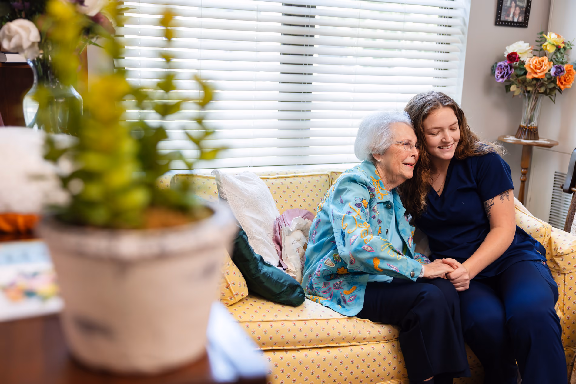 Young caregiver and elderly woman sitting close on a yellow couch, holding hands and smiling.