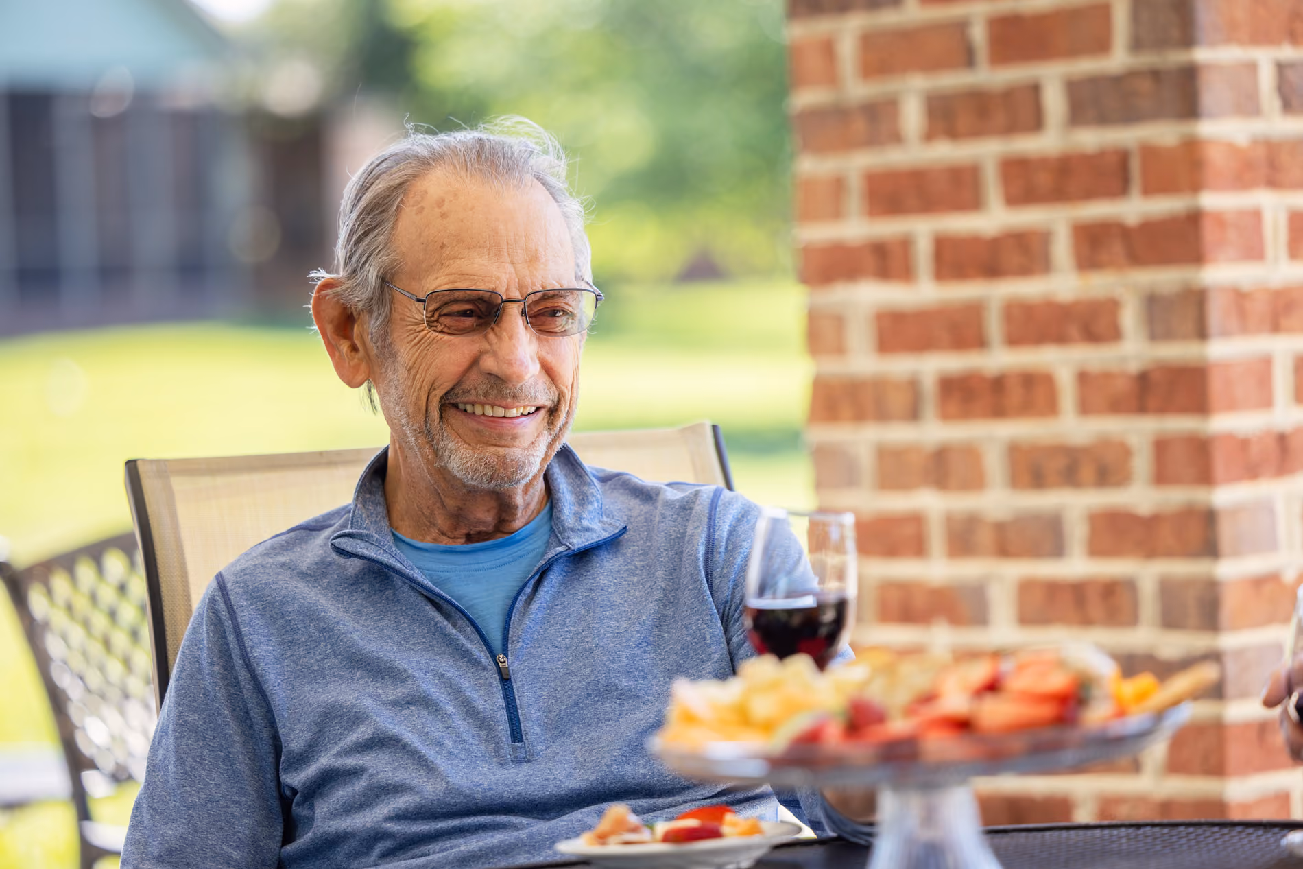 Smiling elderly man in glasses sitting outdoors at a table with a plate of food and a glass of red wine.
