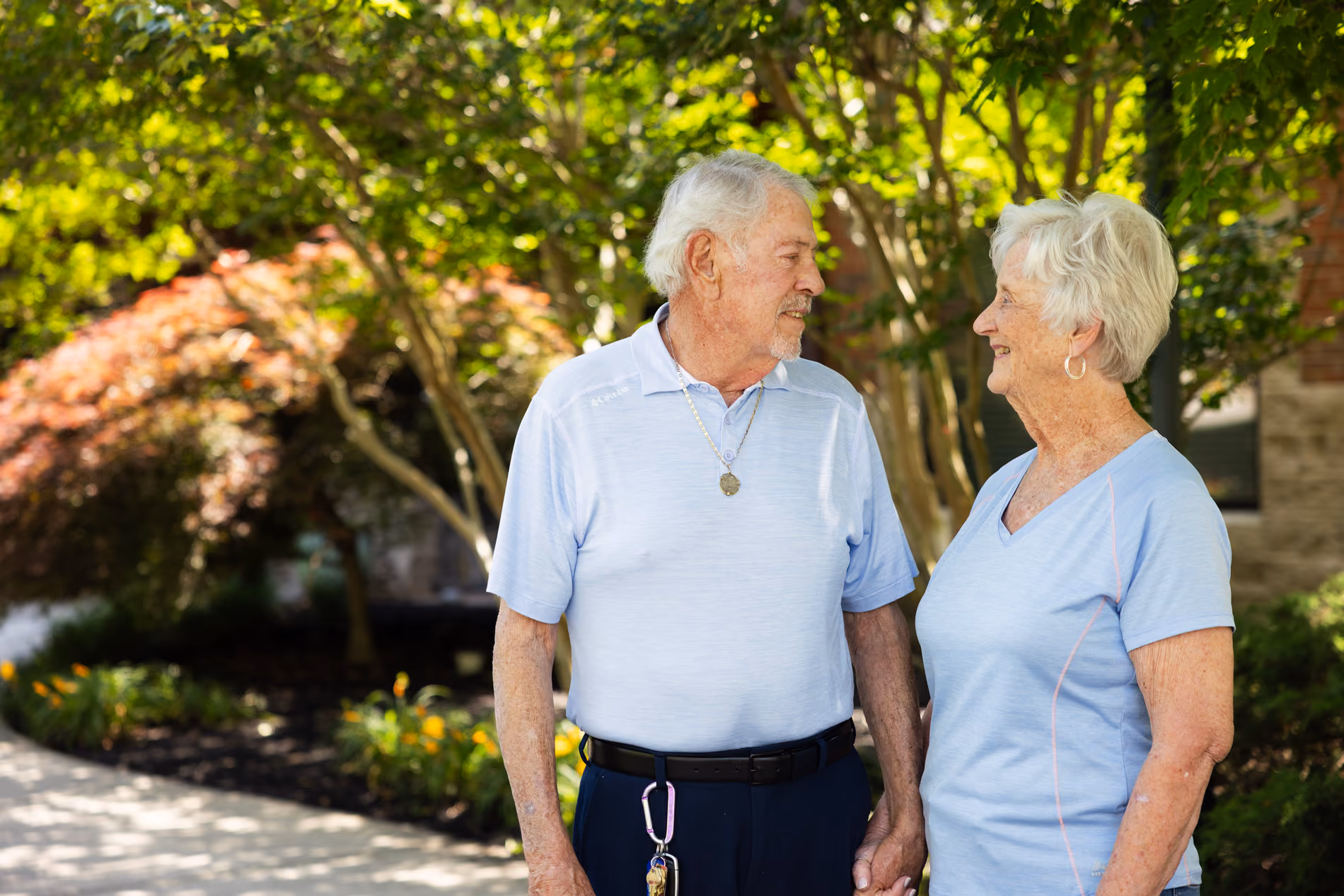Elderly couple holding hands smiling at each other outdoors in a garden with trees and flowers.