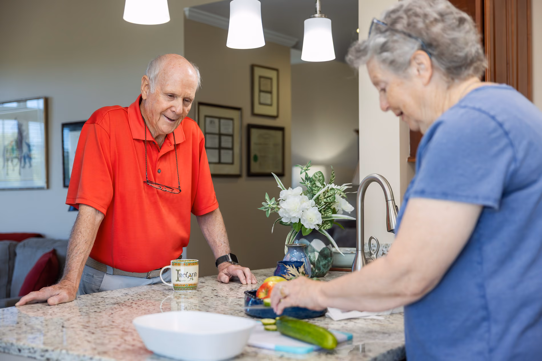 An elderly man in red shirt watches an elderly woman cutting vegetables in a kitchen.