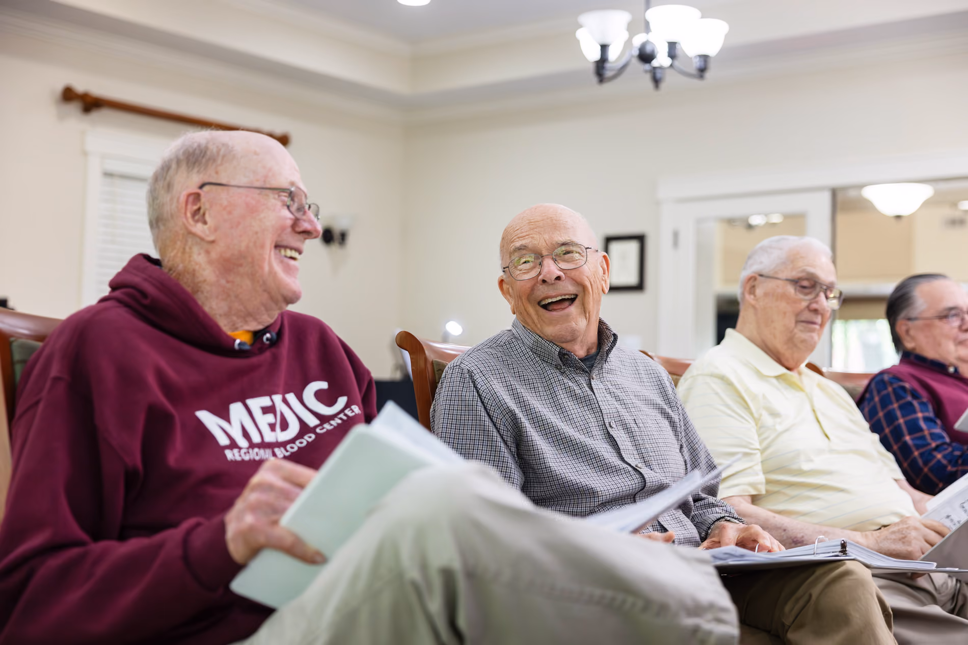 Four elderly men sitting indoors, laughing and holding folders or papers for a group activity.