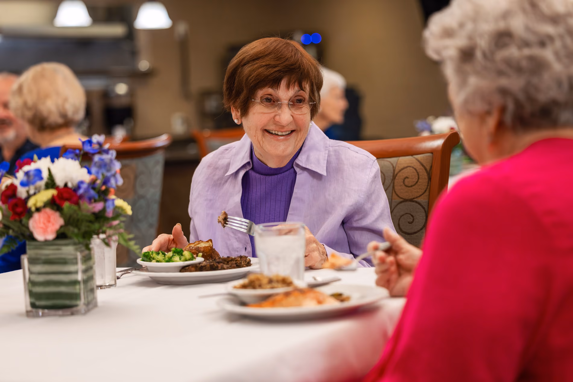 Smiling elderly woman eating a meal and talking with another person at a dining table with flowers.