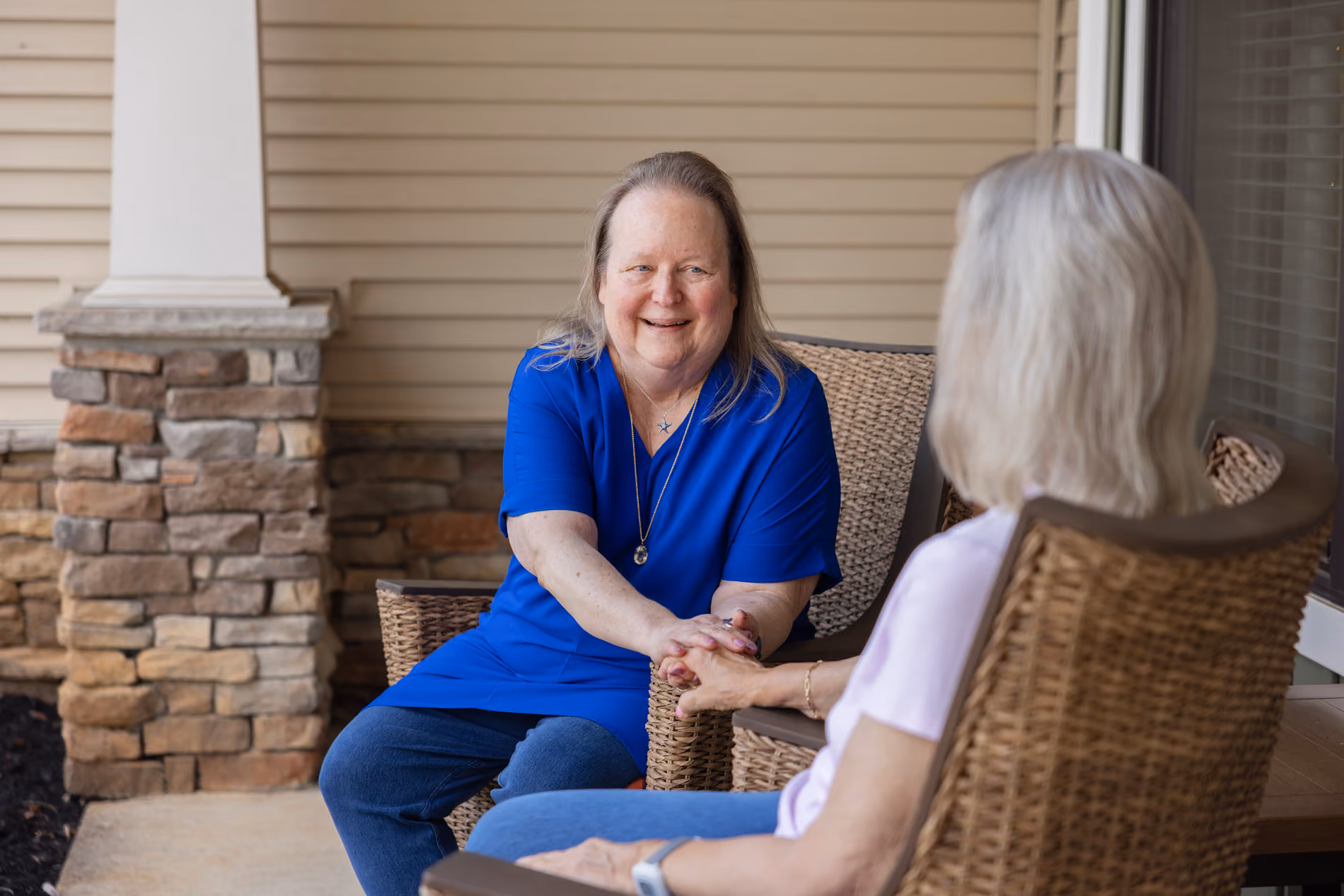 Two elderly women sitting outside on wicker chairs holding hands and smiling at each other.