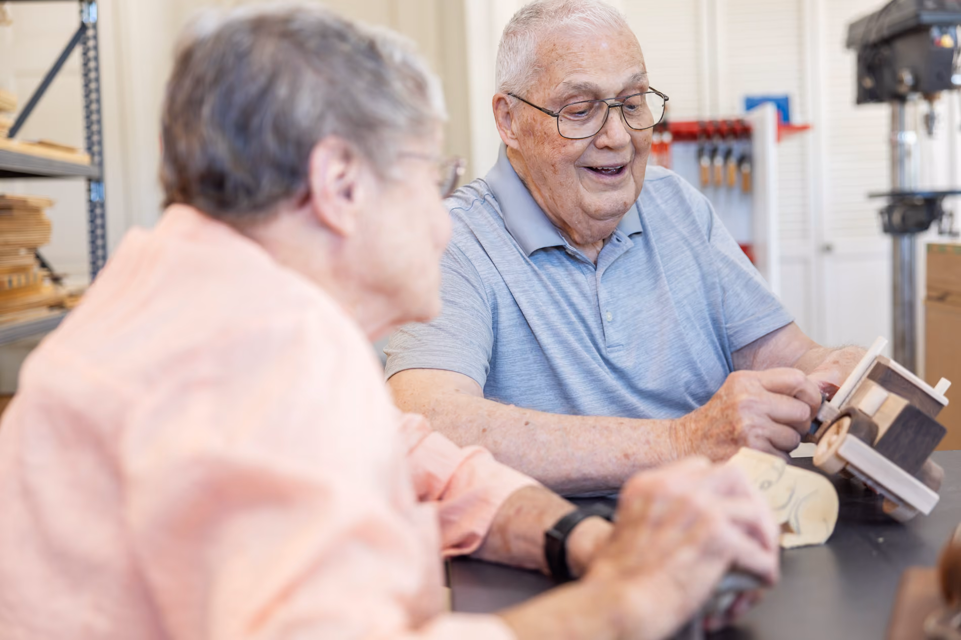 Two elderly people sitting at a table, examining wooden crafts and smiling in a workshop.