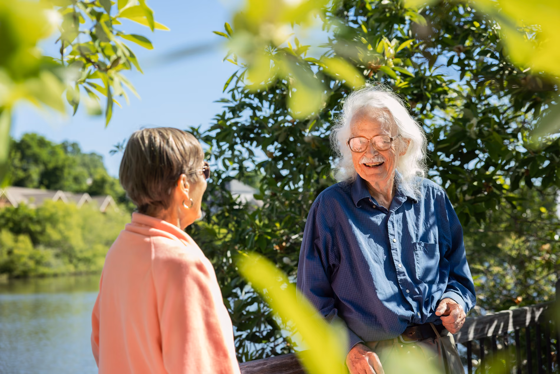 Elderly man and woman smiling and talking outdoors by a lake with trees and houses in the background.