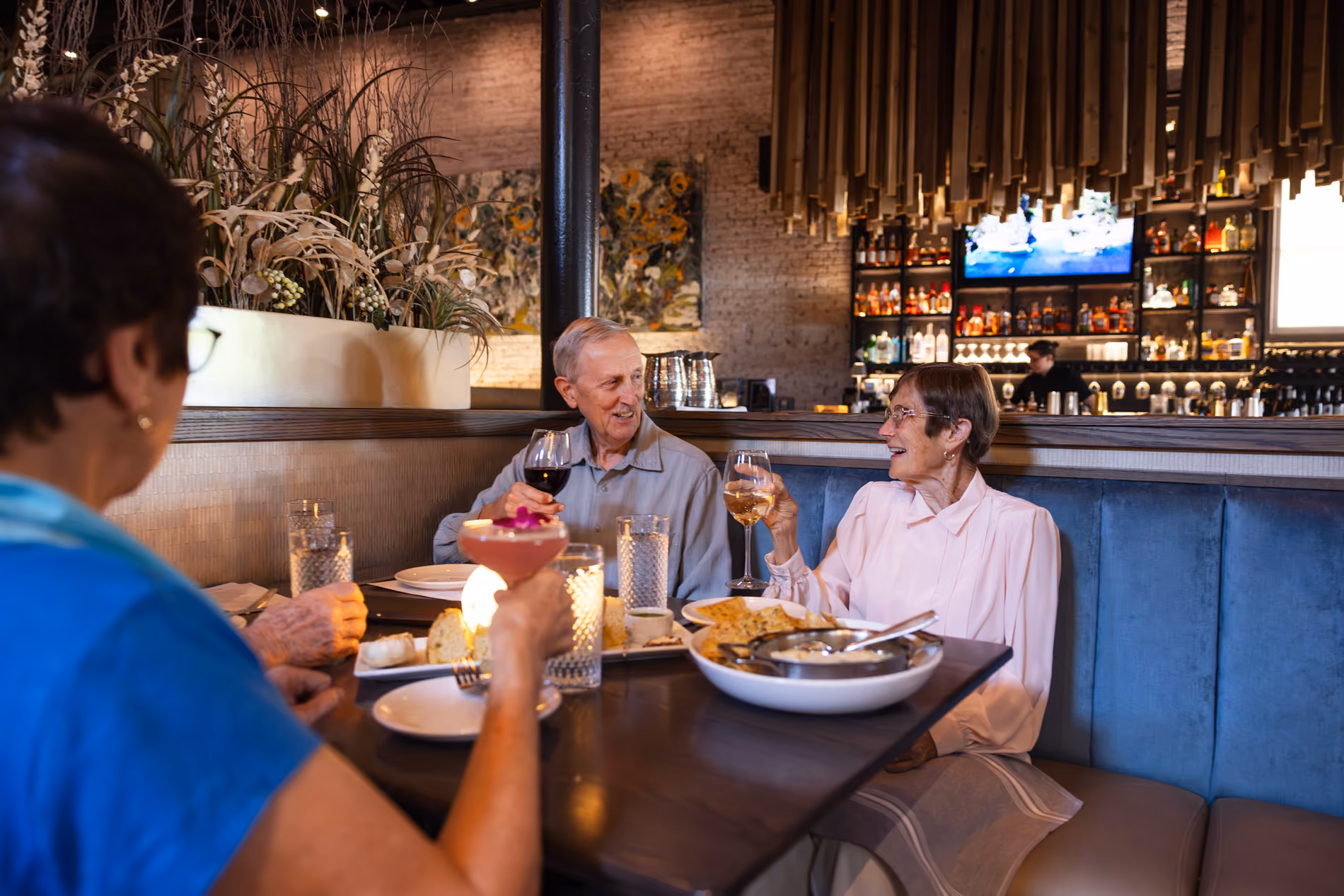 Three elderly people enjoying drinks and food while conversing at a restaurant table booth.