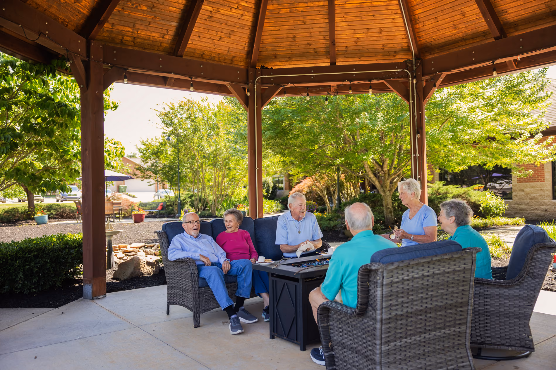 A group of six elderly people sitting and chatting under a wooden gazebo in a garden.