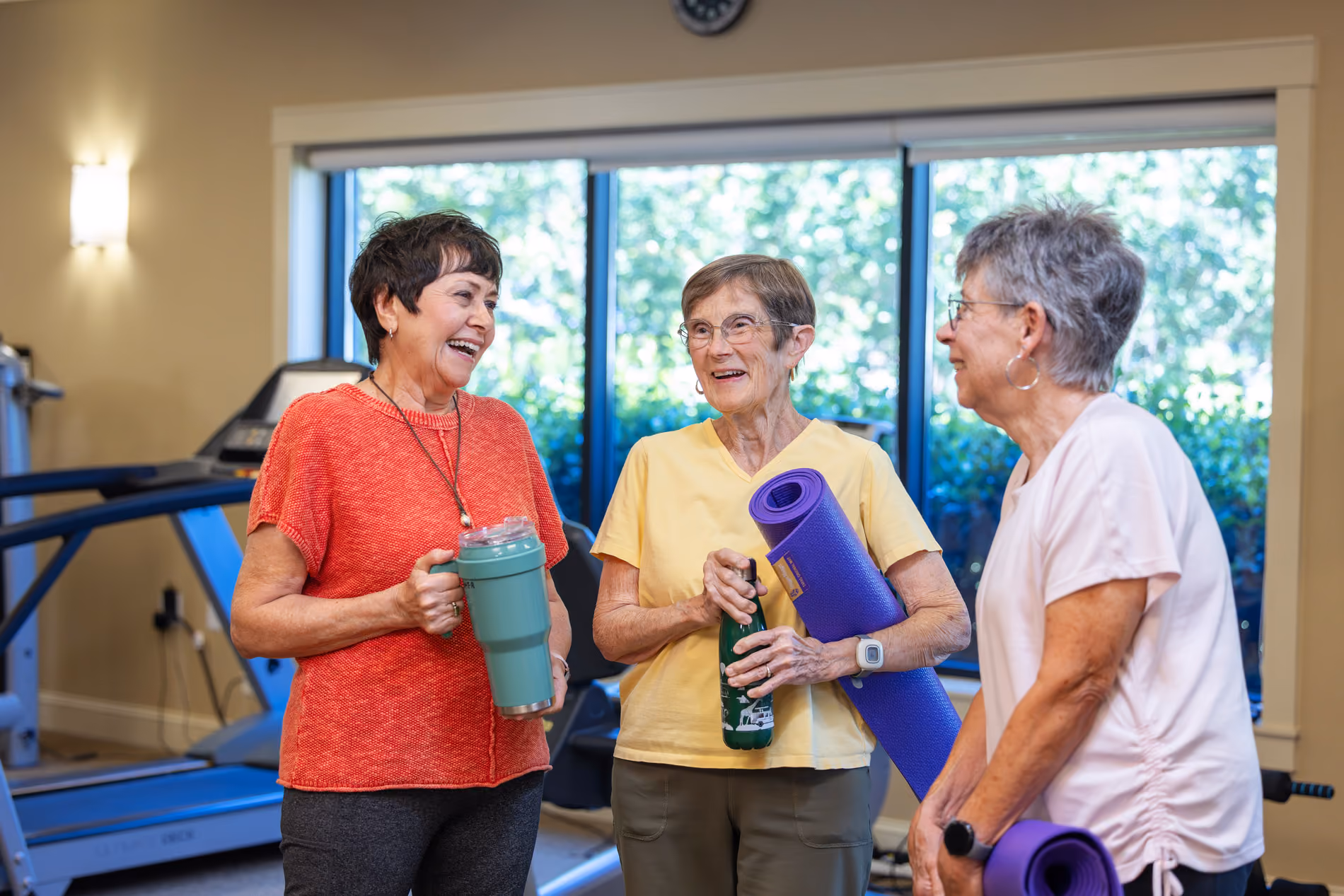 Three older women holding water bottles and yoga mats, chatting and smiling in a gym room.