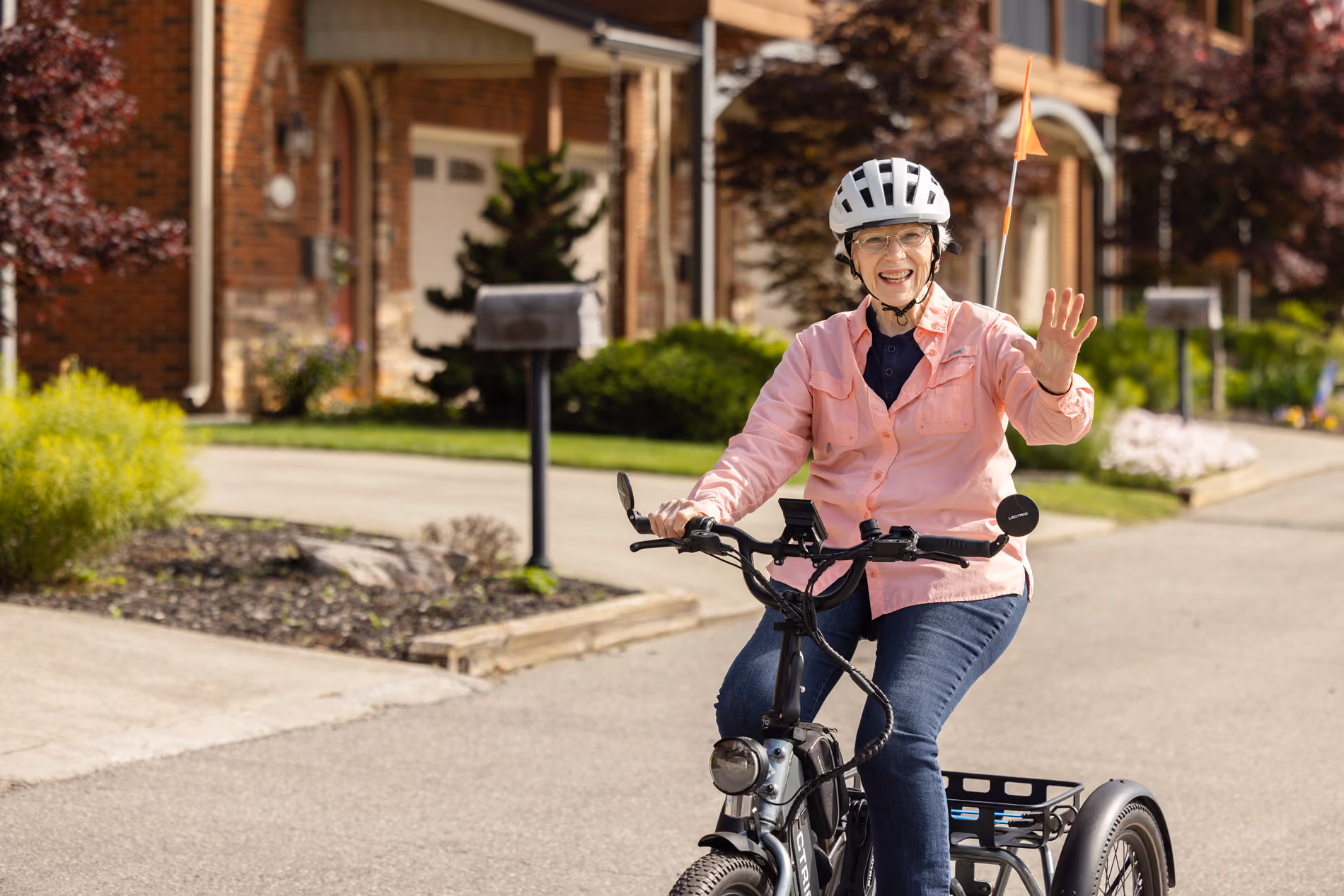 Smiling elderly woman waving while riding a three-wheeled bike on a suburban street.