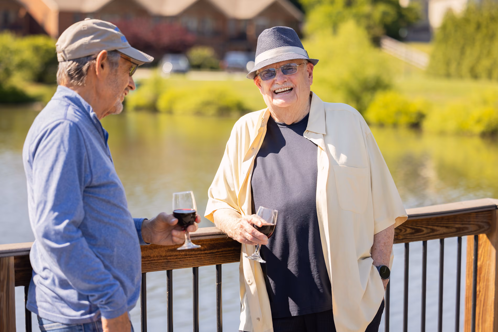 Two older men smiling and holding glasses of red wine on a wooden deck by a lake.